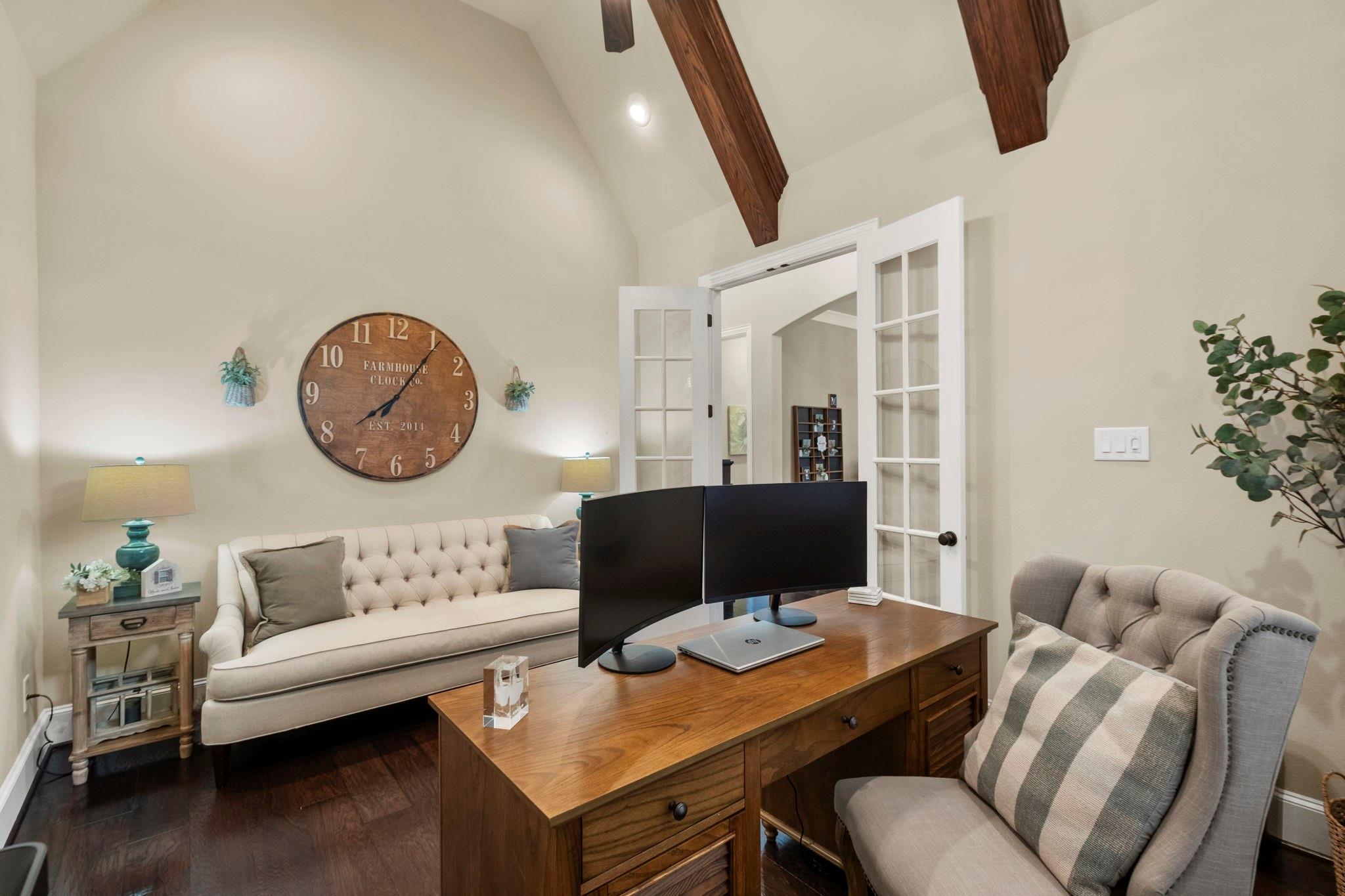 Home office with a wooden desk and dual monitors, tufted sofa, and exposed ceiling beams.