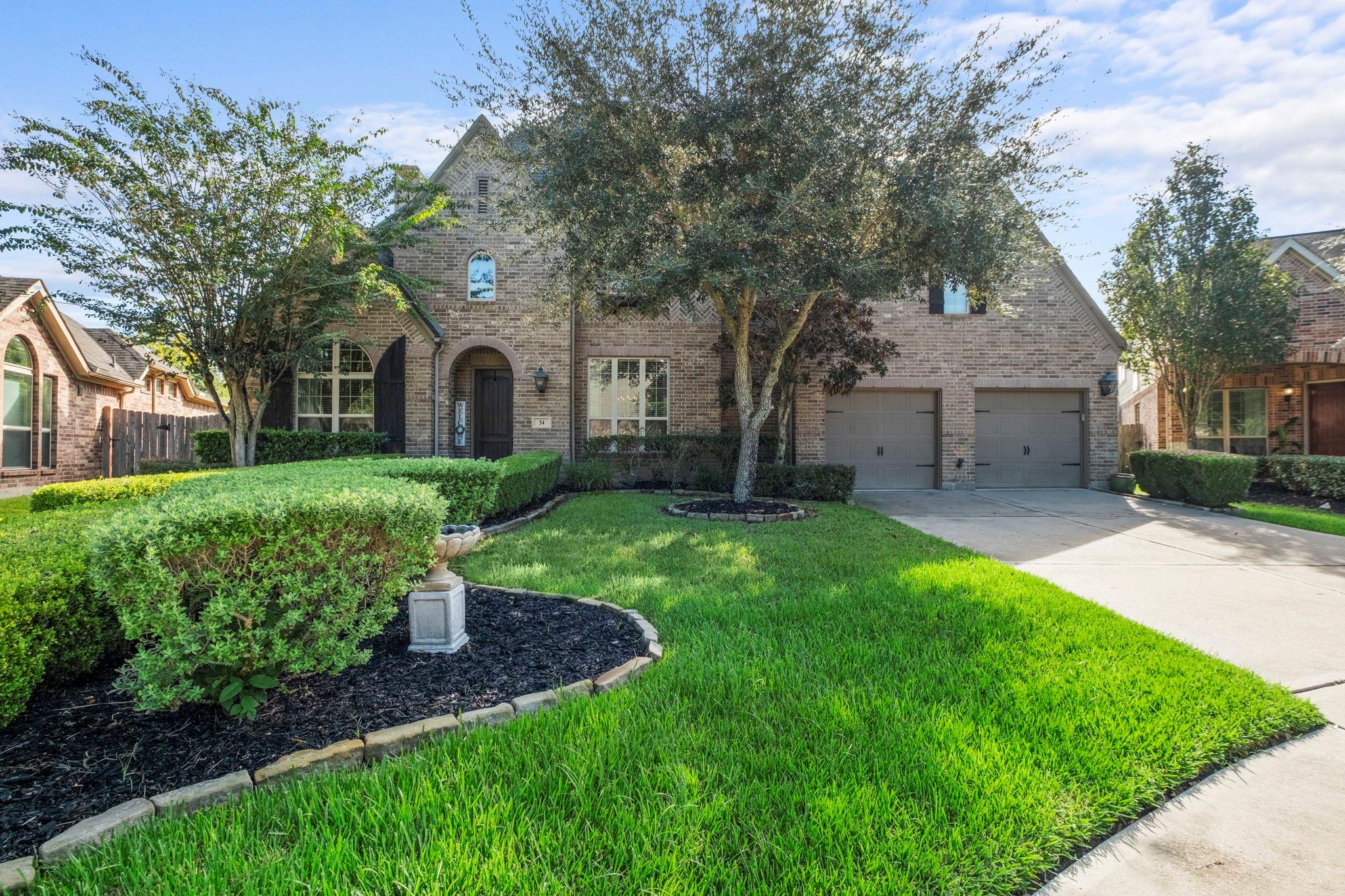 Brick house with arched entry, two-car garage, mature trees, and landscaped lawn.