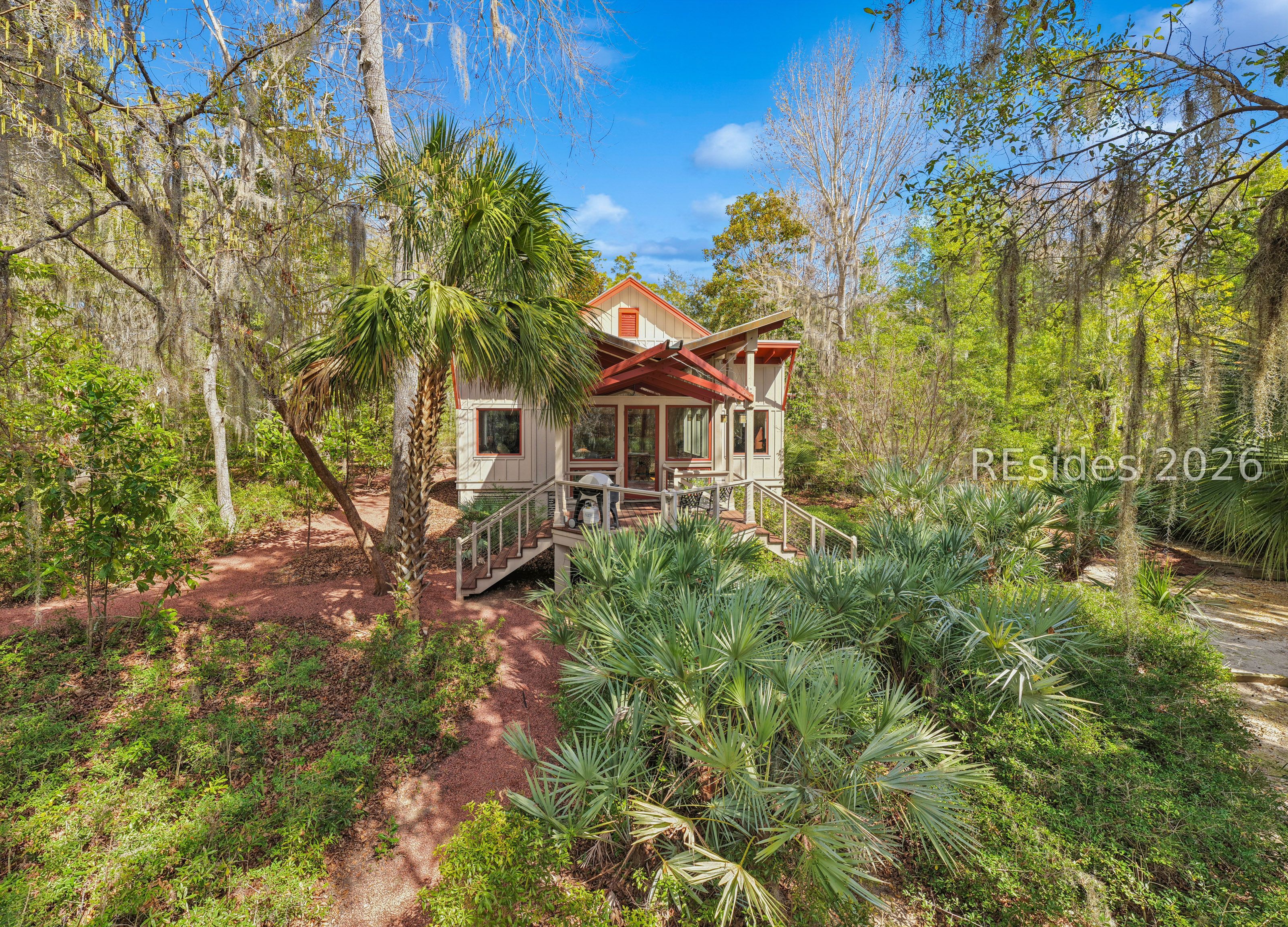 A raised light-sided home with red trim and a deck sits among palms and moss-draped trees.