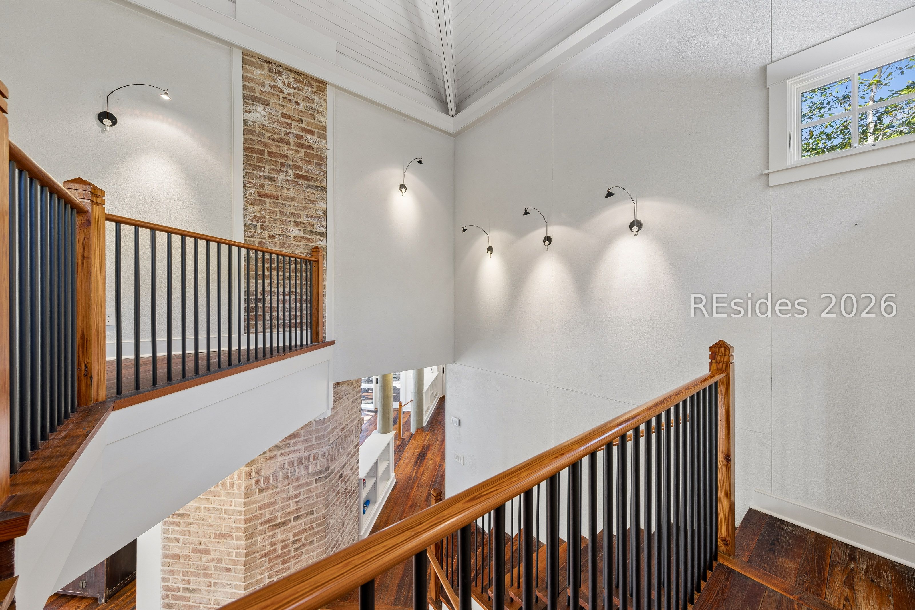 Open staircase with wood handrails and black balusters beside a brick accent wall in a vaulted stairwell.