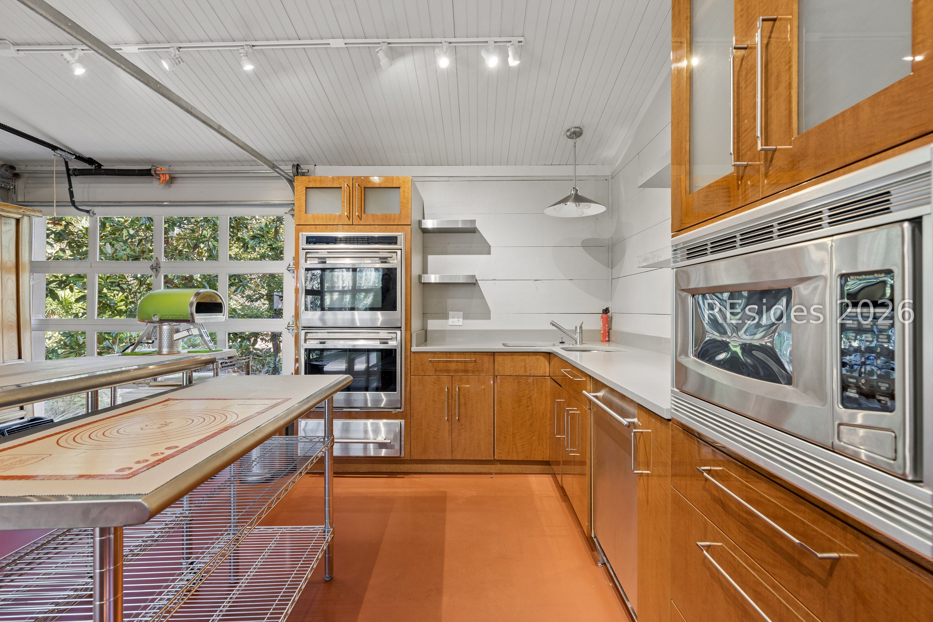 Kitchen with warm wood cabinets, stainless steel appliances, and a metal worktable beside leafy windows.