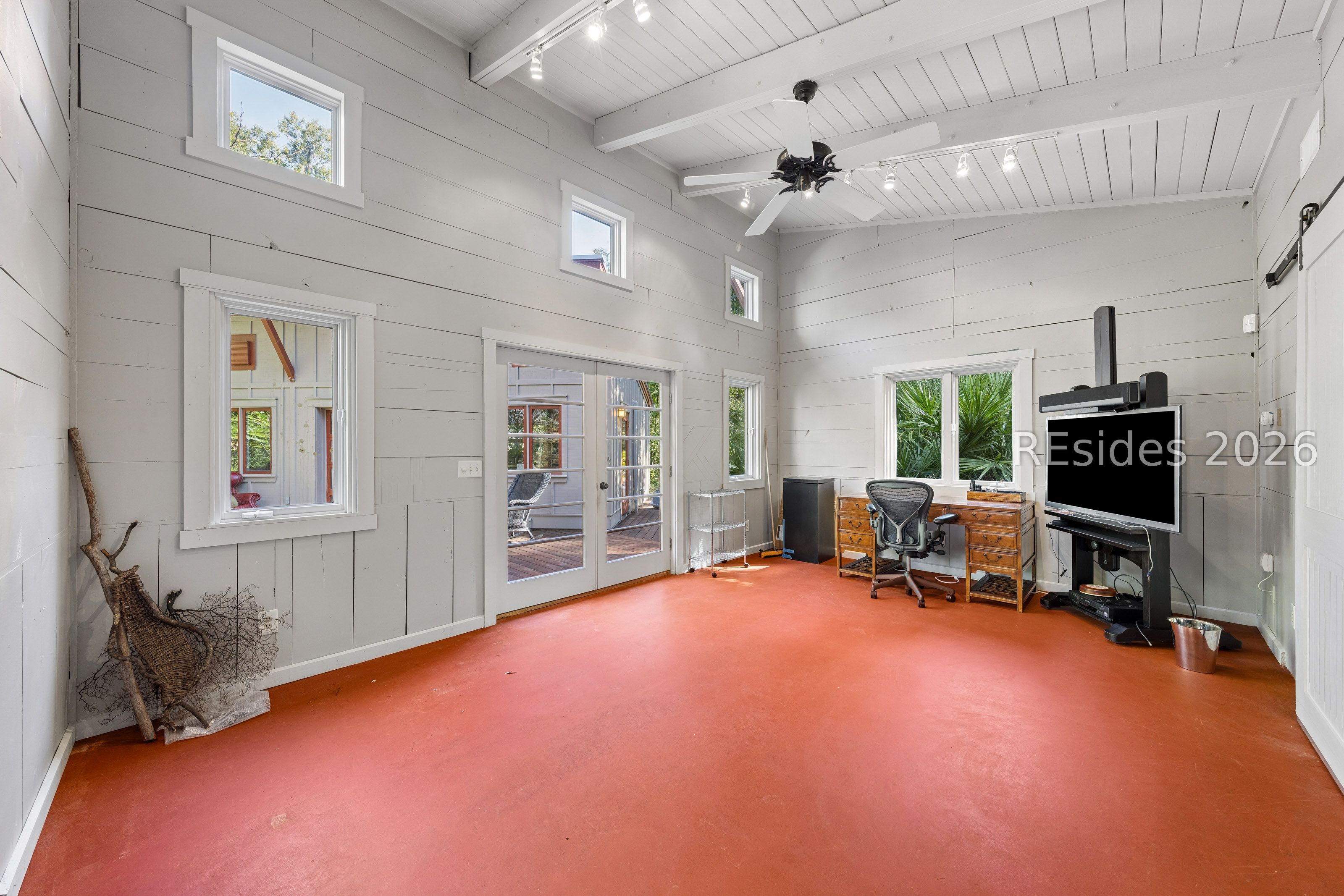 Bright office with a red-orange floor, white shiplap walls, French doors, and a high beamed ceiling.