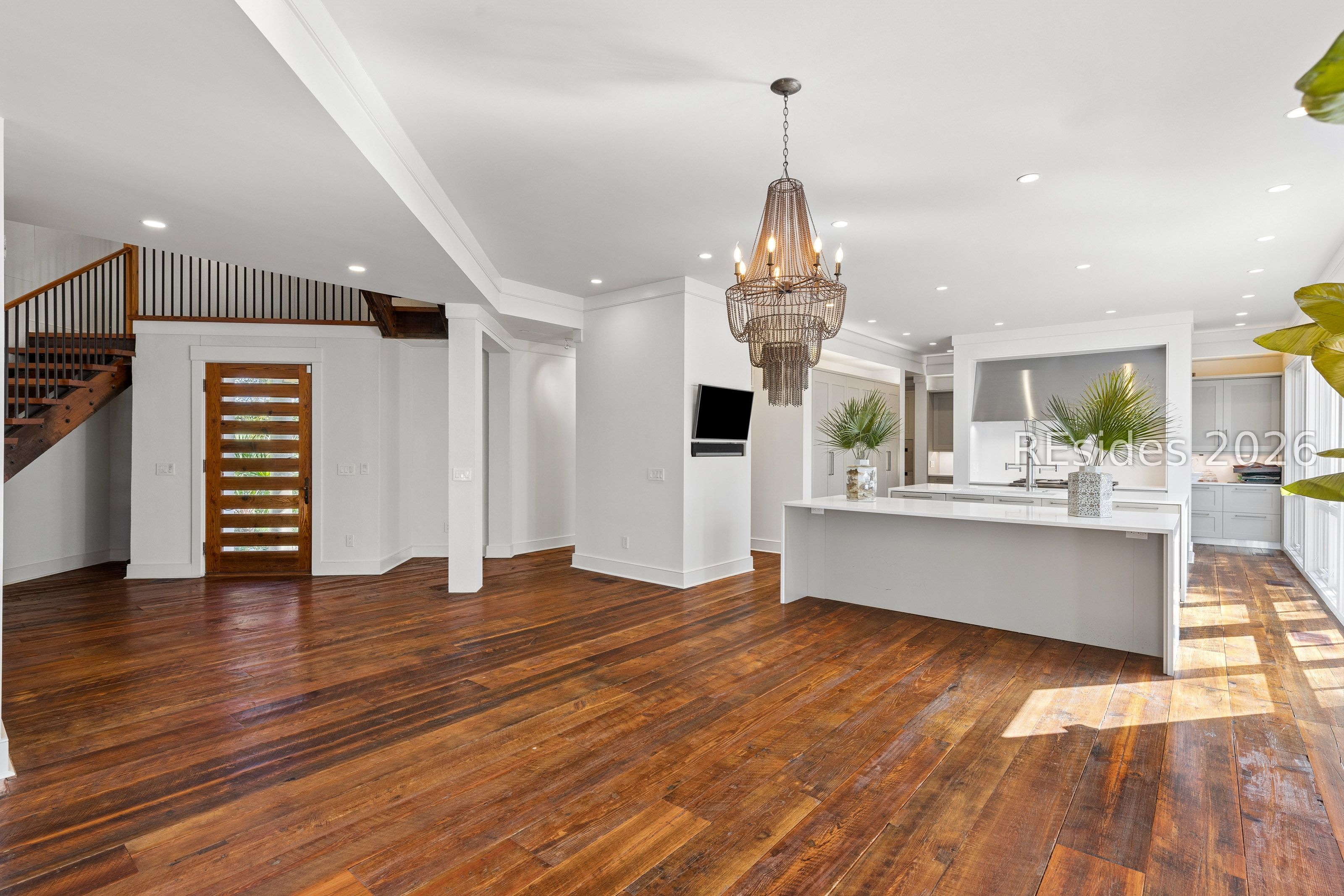 Open-plan kitchen with a white island, woven chandelier, hardwood floors, and a wooden staircase.