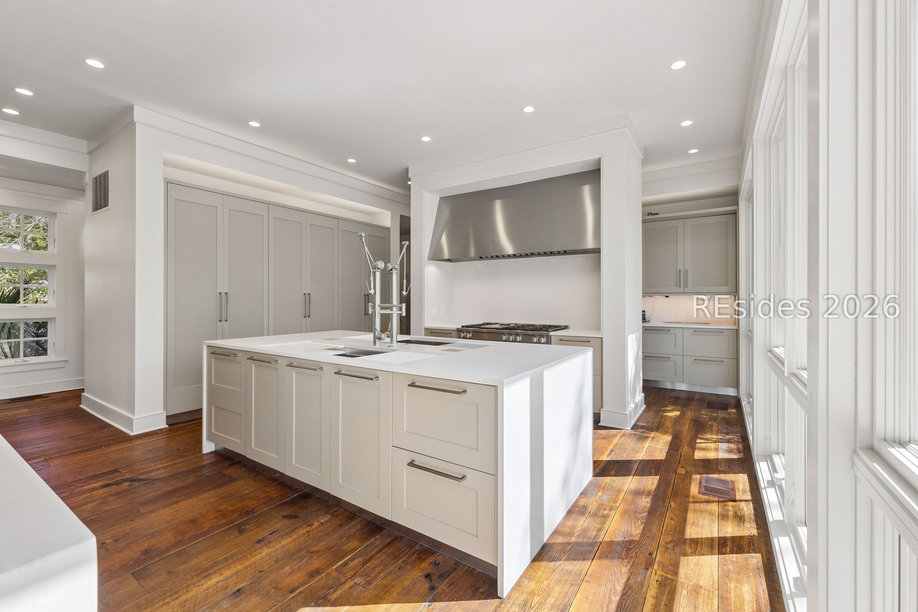 Bright kitchen with a large white island, gray cabinetry, stainless-steel range hood, and sunlit wood floors.