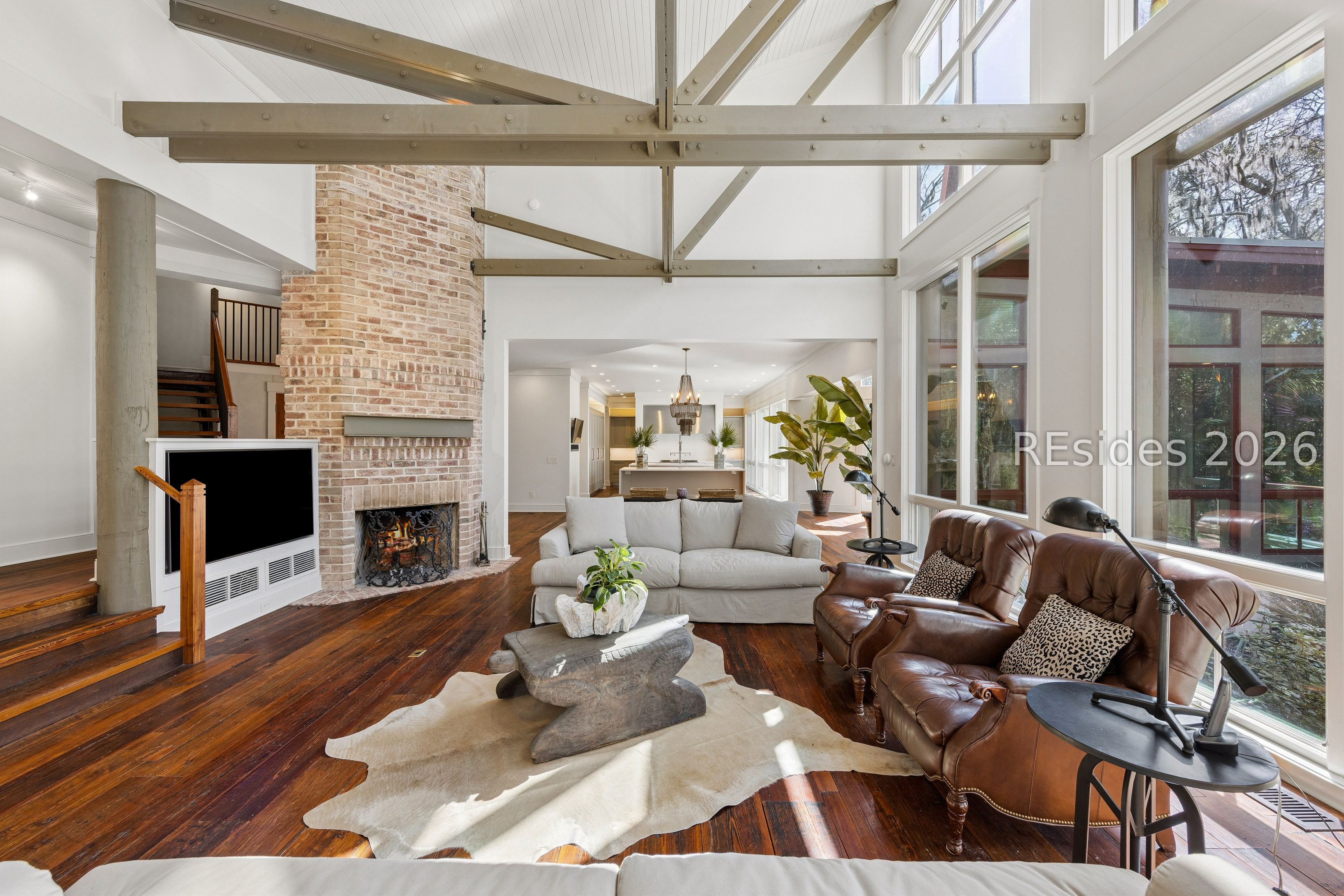 Living room with vaulted beams, a brick fireplace, leather chairs, and tall windows.