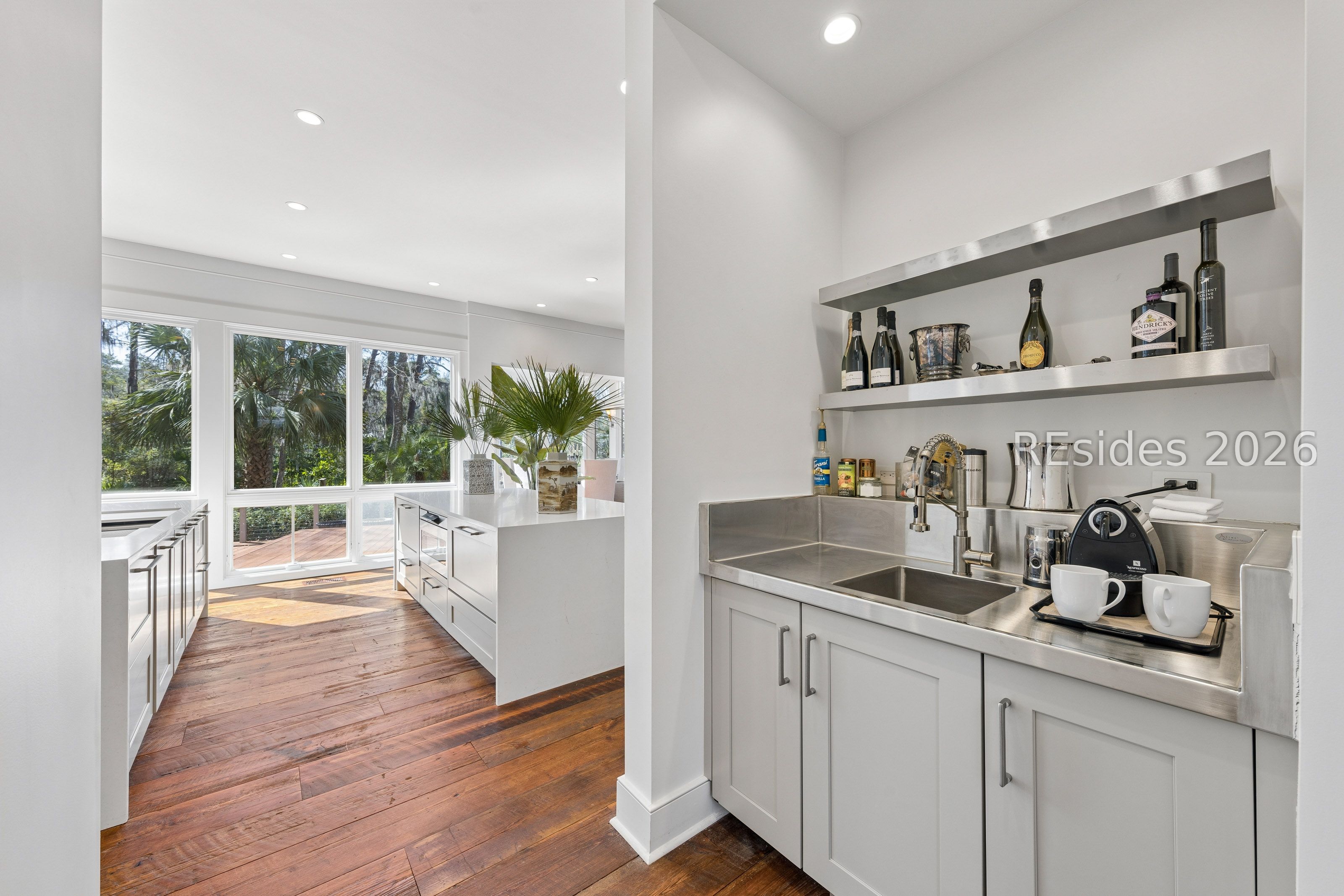 Bright kitchen with stainless-steel sink, open shelves, wood floors, and large windows overlooking palms.