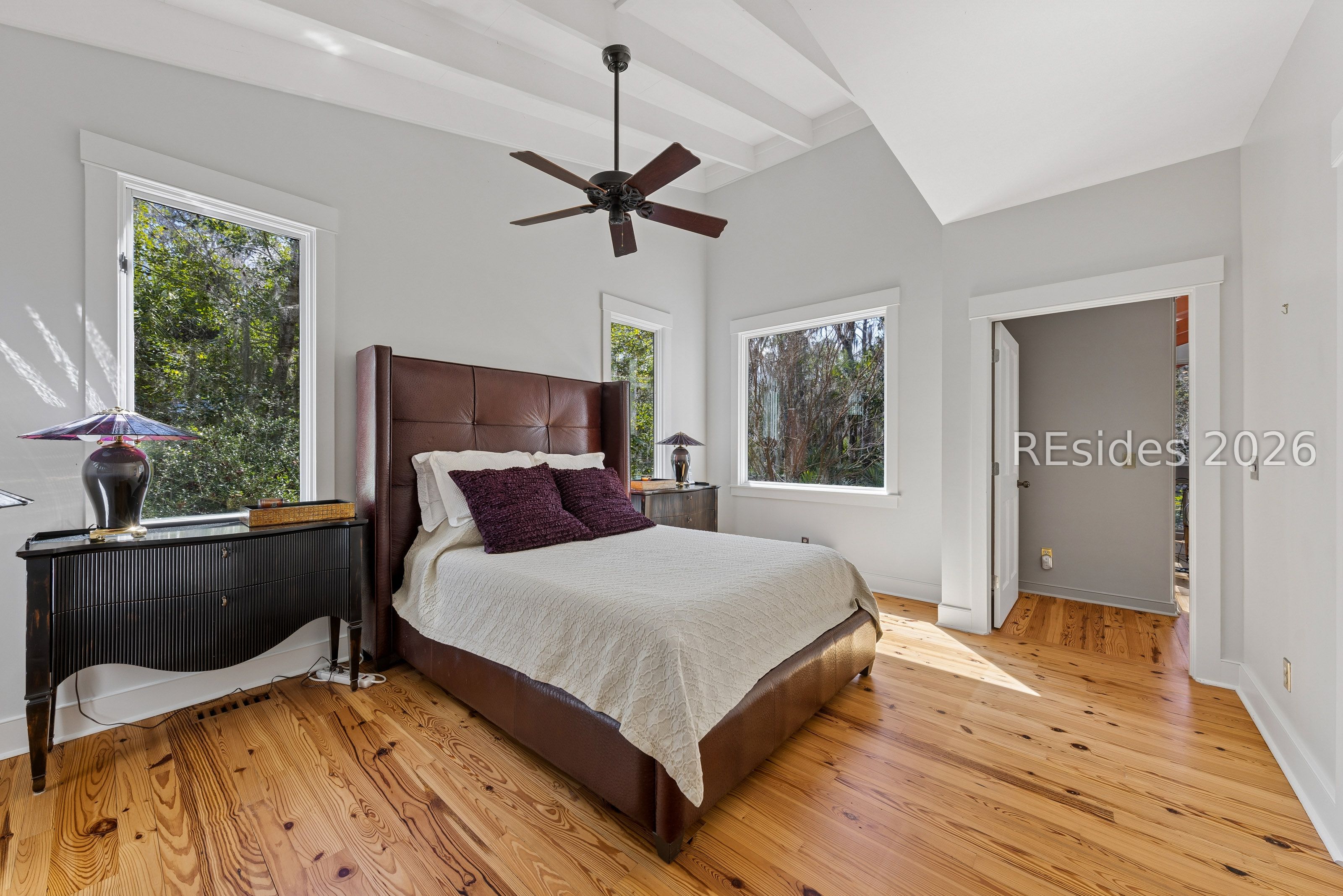 Bedroom with a tall leather bed, exposed ceiling beams, warm wood floors, and windows facing trees.