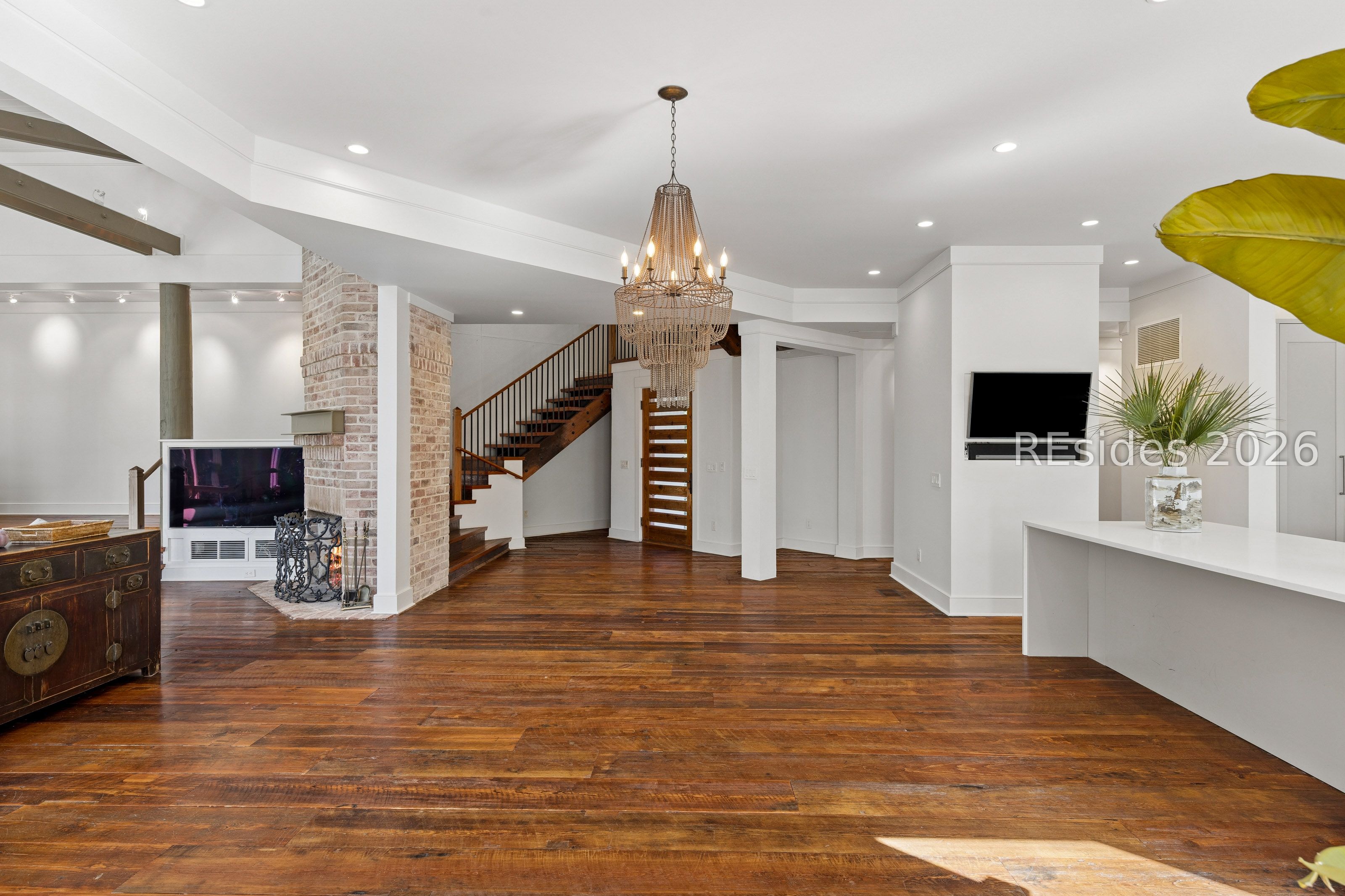 Open living area with dark wood floors, brick fireplace, and a chandelier beside the staircase.