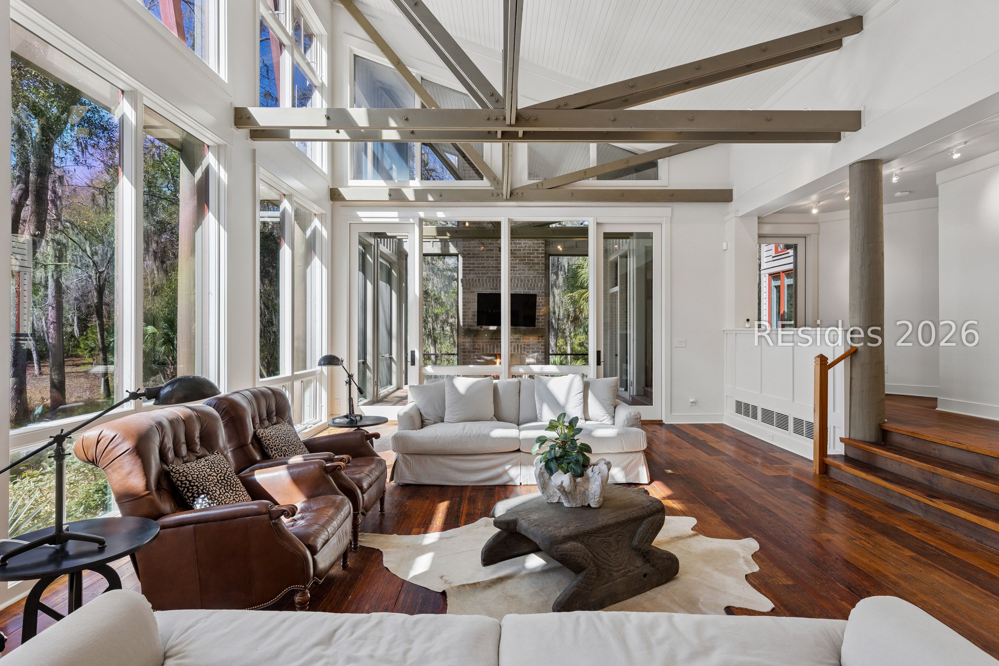 Living room with tall wooded-view windows, exposed beams, hardwood floors, and leather and white sofas.