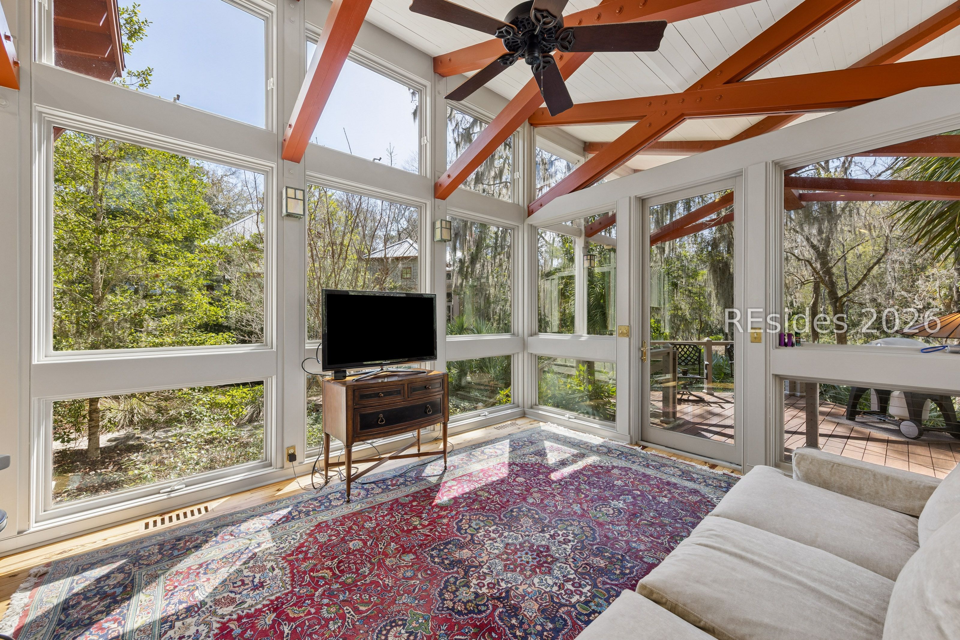 Sunroom with tall windows, exposed wood beams, and a patterned rug overlooking trees and a deck.