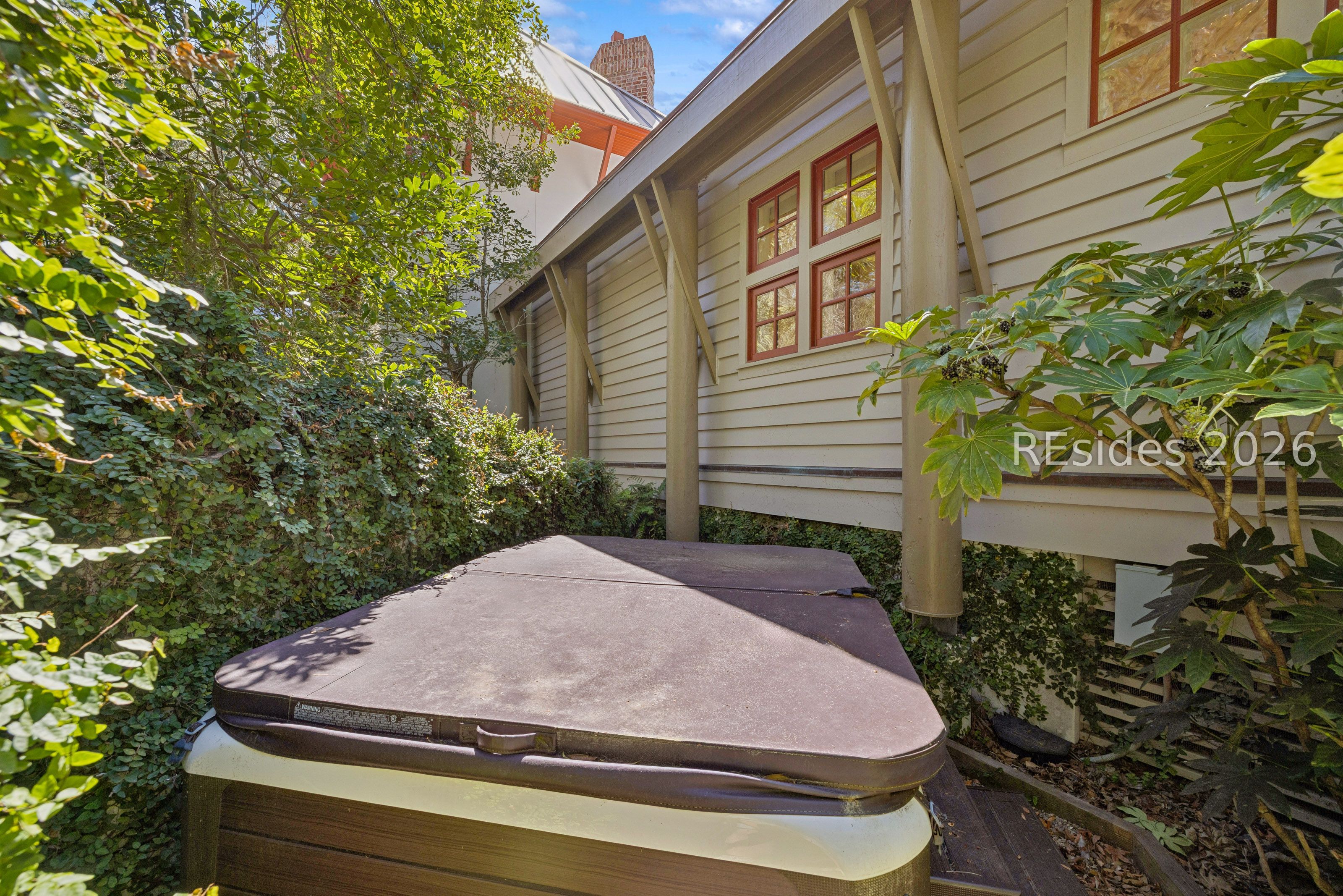 Outdoor hot tub beside a leafy, raised home exterior with red-trimmed windows.