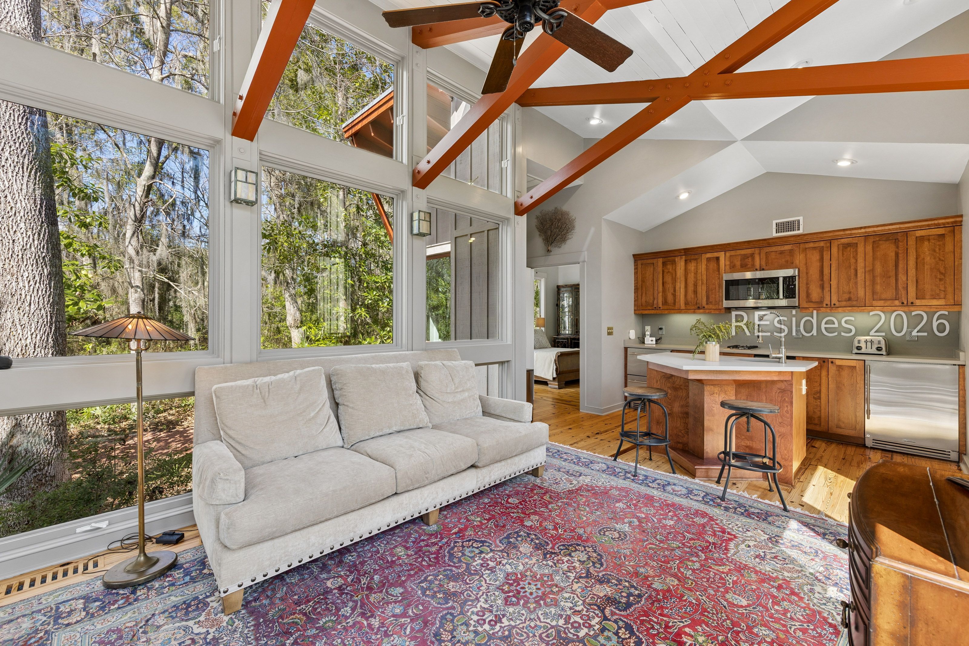 Living room with vaulted exposed beams, tall windows overlooking trees, and an open kitchen.