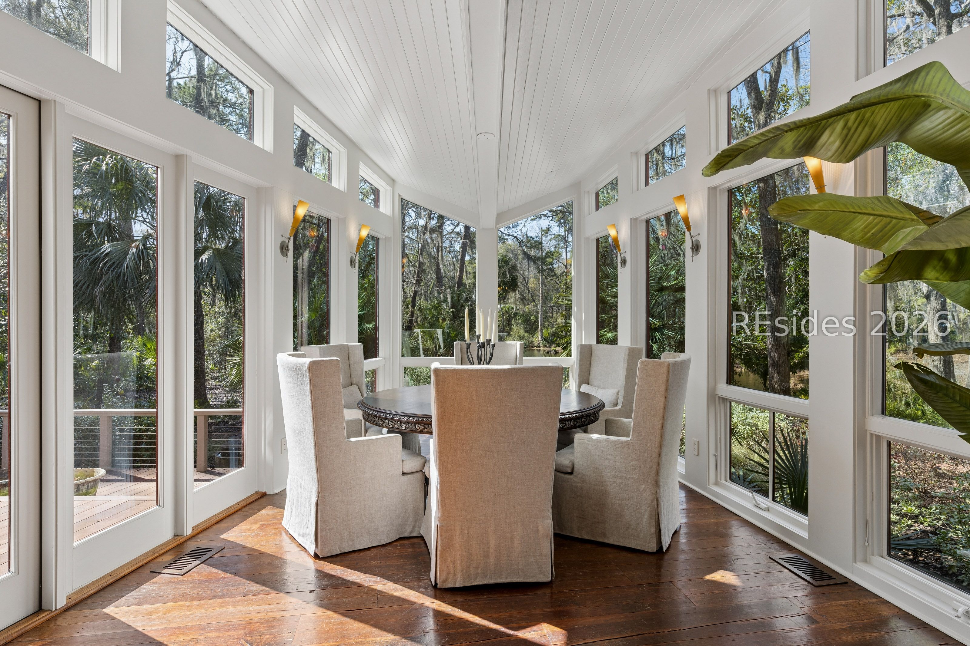Sunlit dining room with a round table, upholstered chairs, and tall woodland-facing windows.