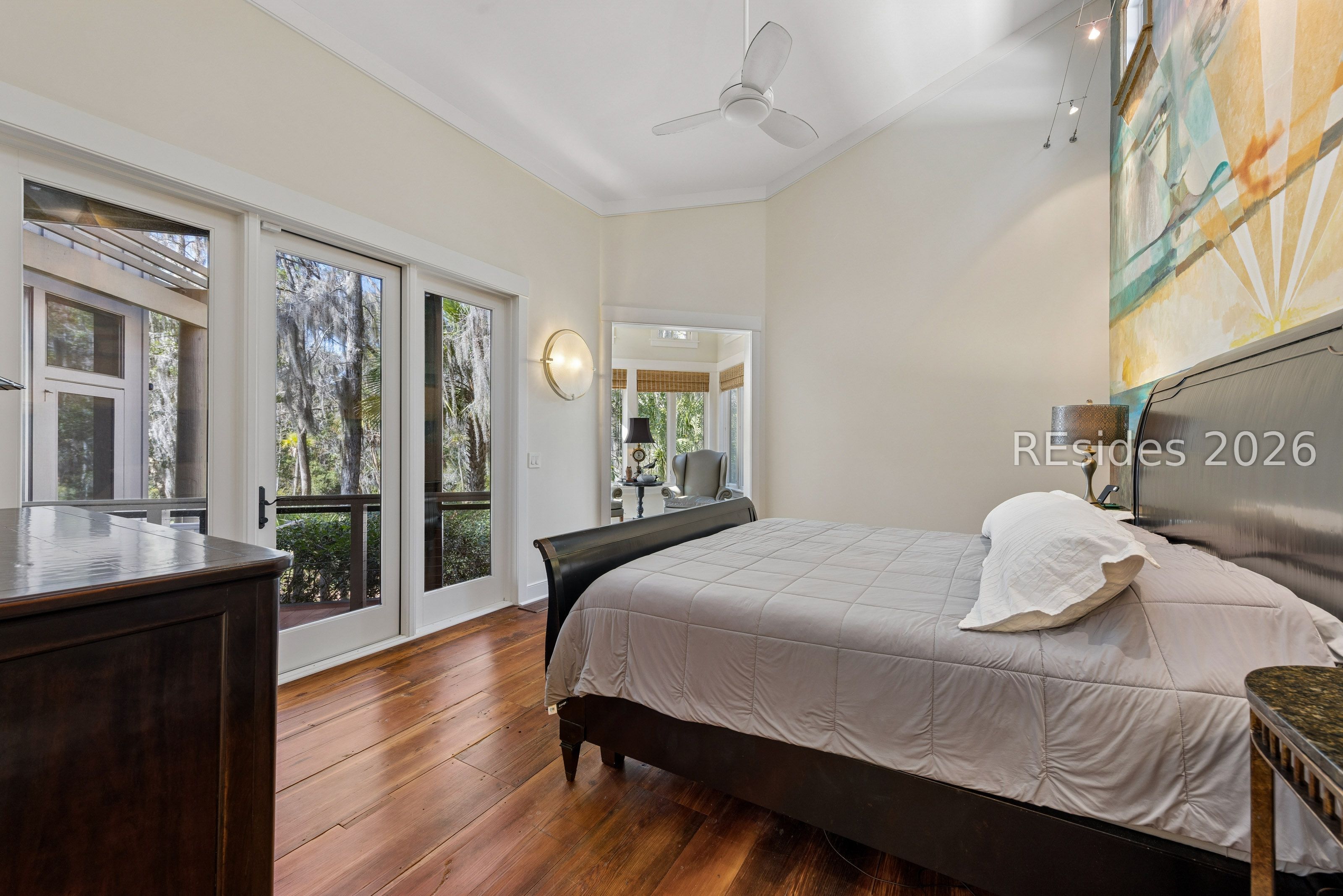 Bedroom with a gray-covered bed, hardwood floors, and glass doors overlooking trees.