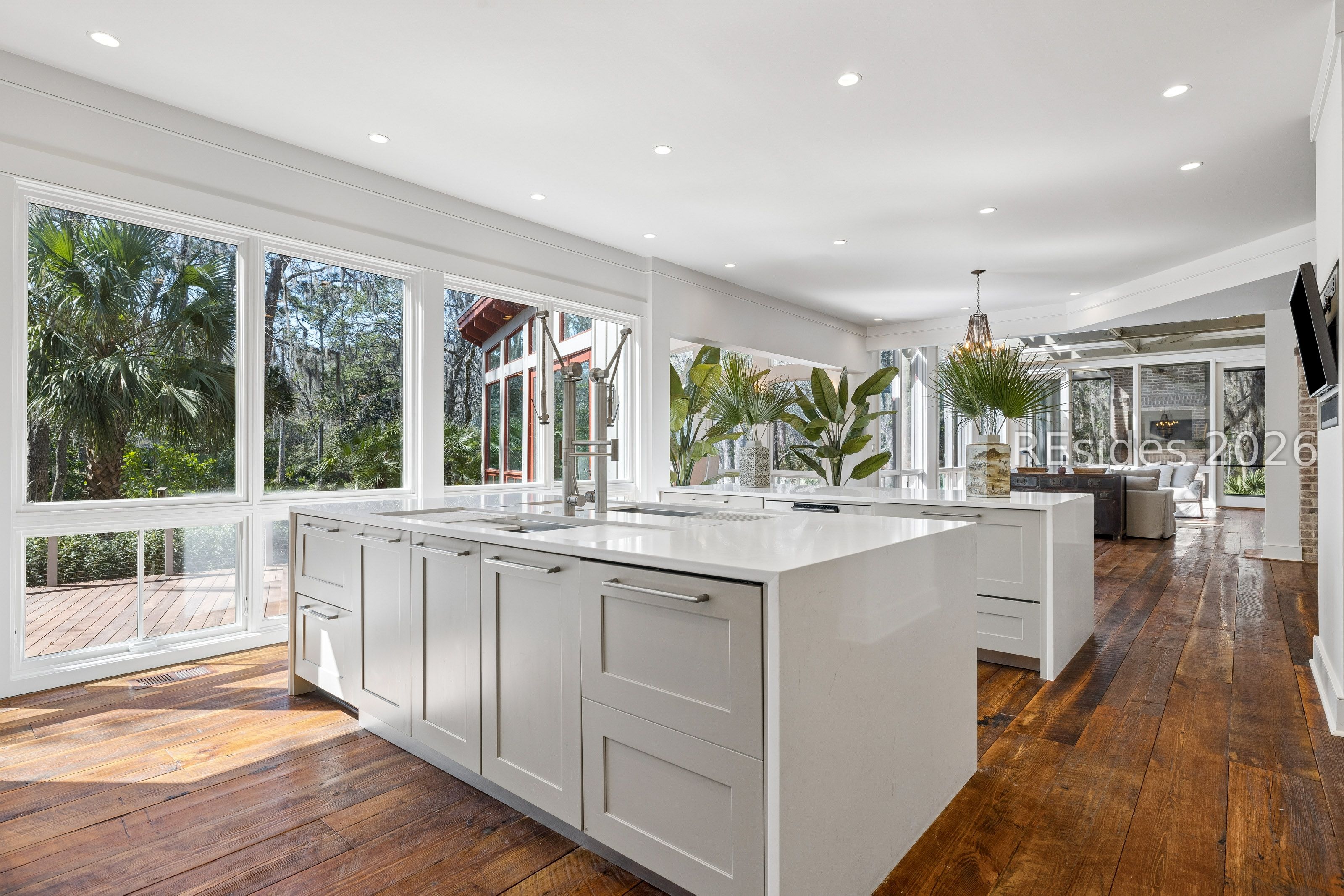 Bright white kitchen with a large island, wide windows overlooking greenery, and dark wood floors.