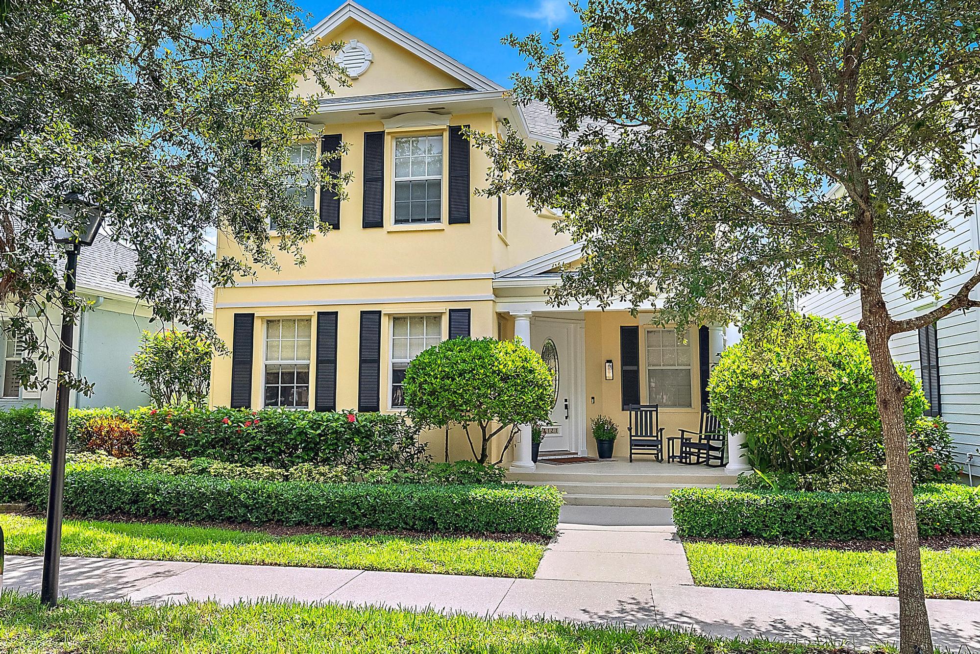 Pale yellow two-story home with black shutters, a columned porch, and a leafy landscaped front yard.