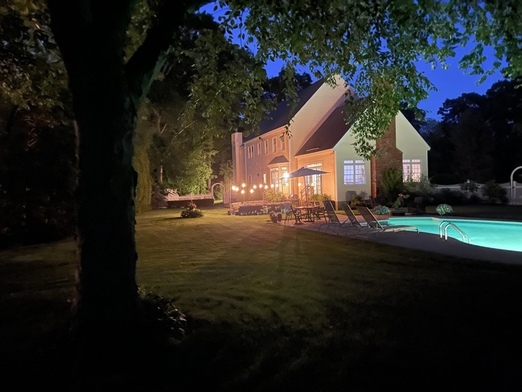 Backyard of a large house at night, with a string-lit patio and illuminated swimming pool.