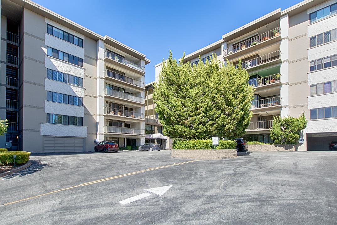Multi-story apartment complex with balconies and a large evergreen tree in the courtyard.