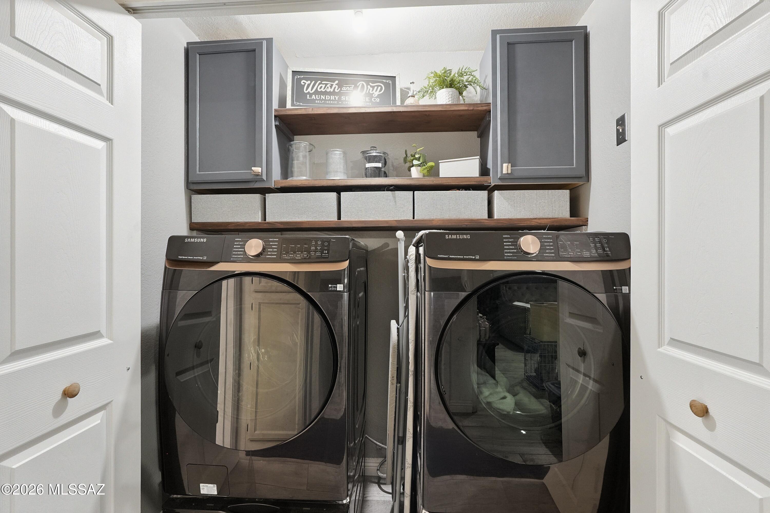 Laundry room with dark Samsung washer and dryer beneath wood shelves and gray cabinets.
