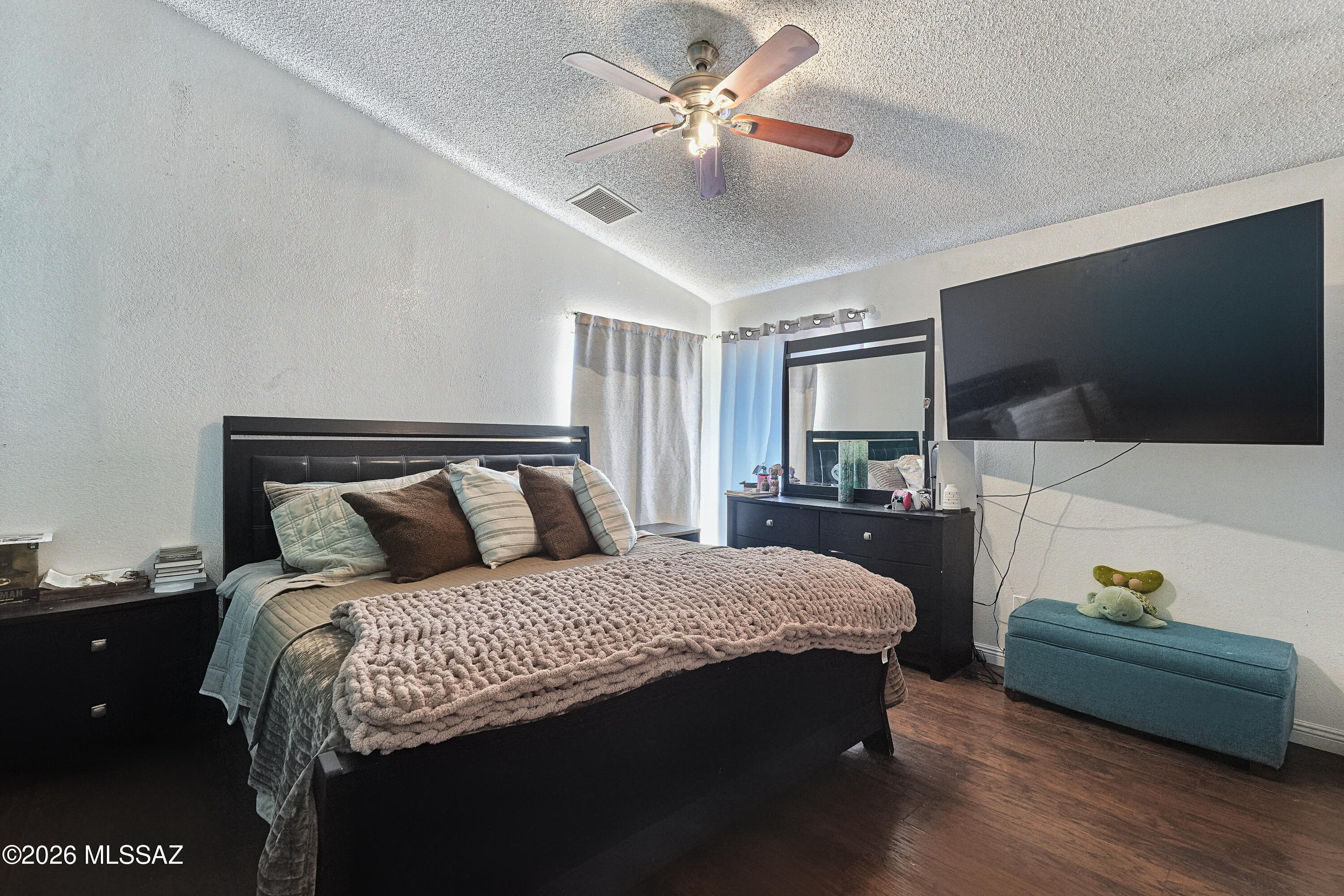 Furnished bedroom with a dark bed, chunky knit blanket, wall-mounted TV, and blue bench.