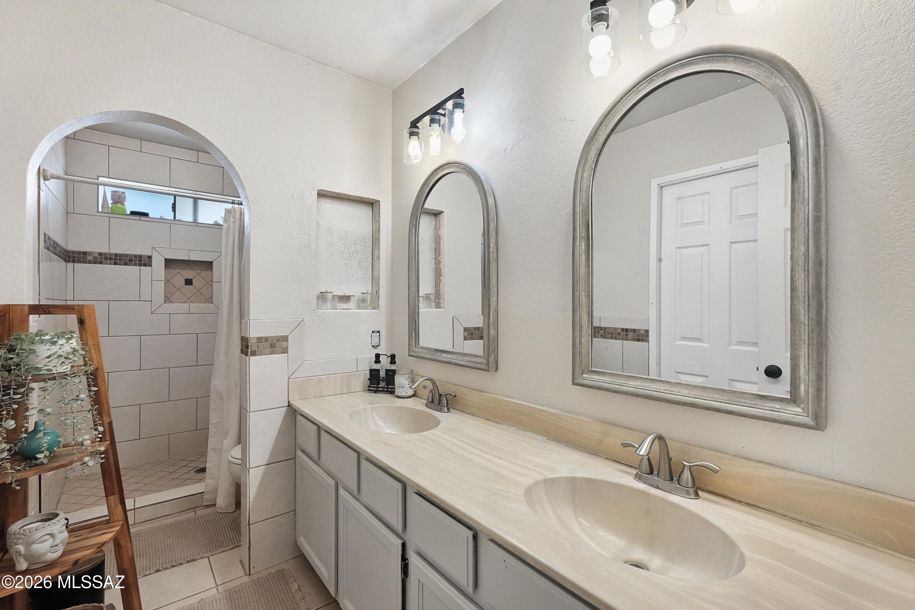 Bathroom with a long double vanity, arched mirrors, and a tiled shower with an accent band.