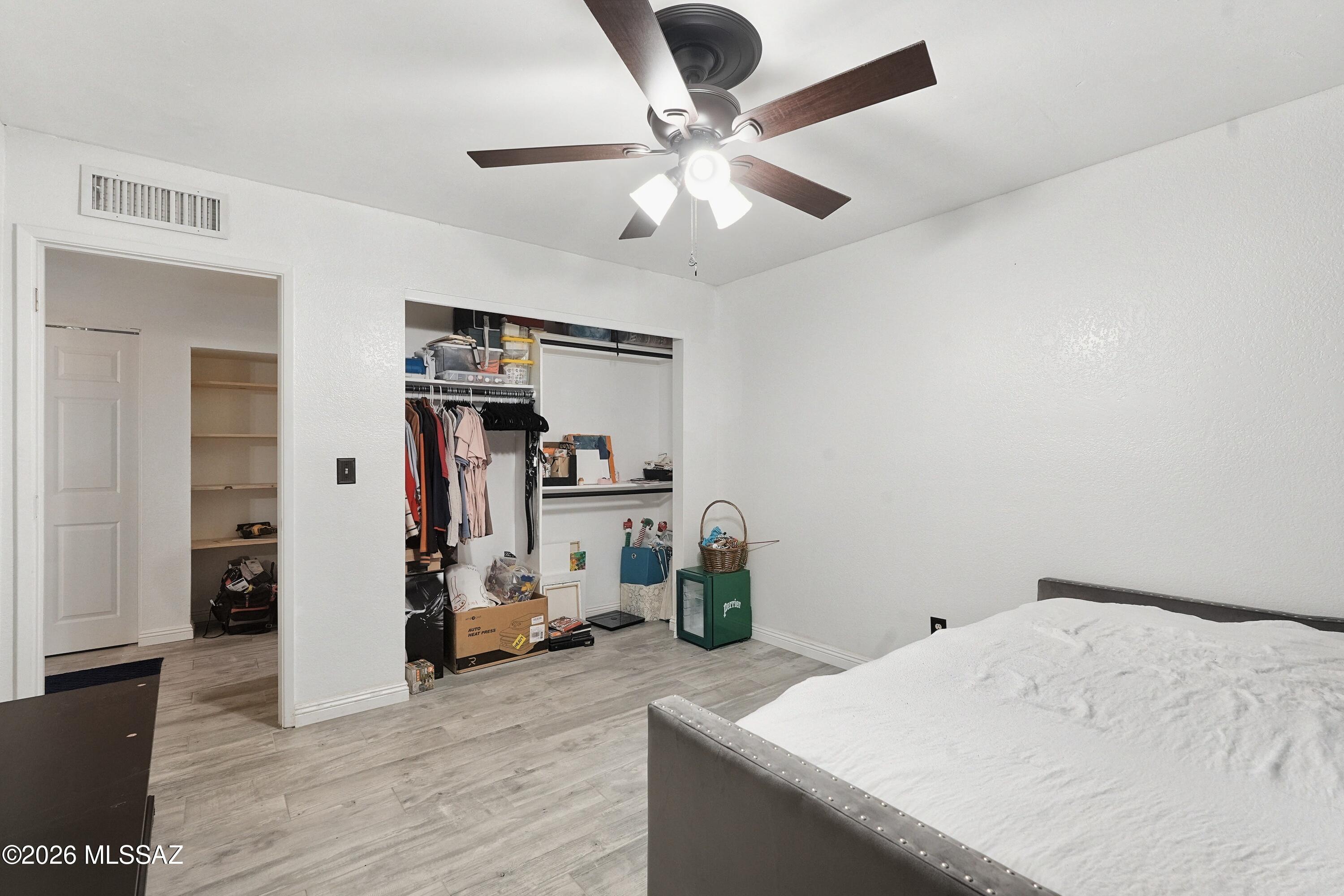 Bedroom with a gray bed and ceiling fan, showing open closets filled with clothing and storage items.