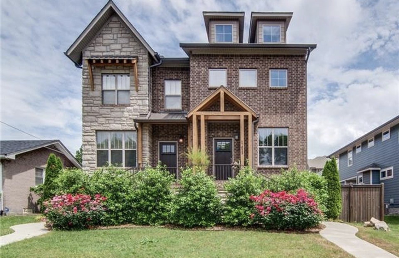 Two-story brick and stone home with a timber porch and flowering shrubs along the front garden.