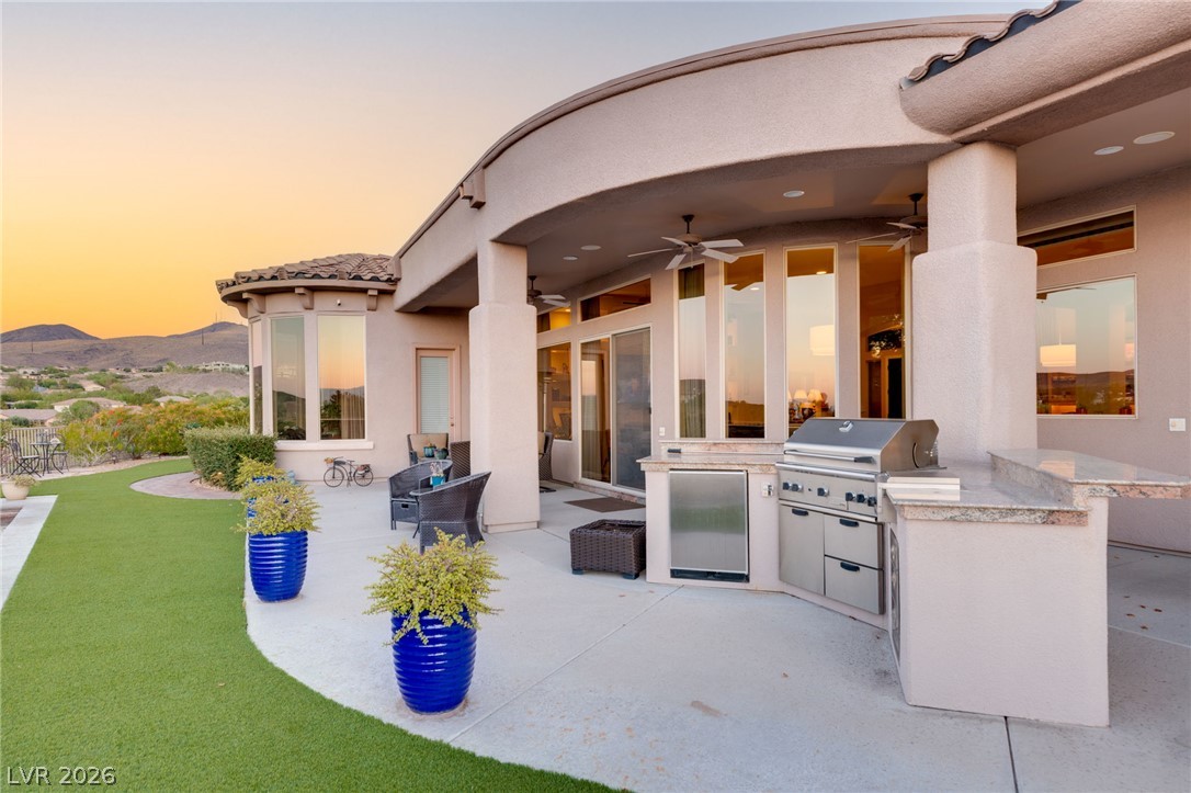 Covered rear patio with an outdoor kitchen and grill, artificial lawn, and desert hills at sunset.