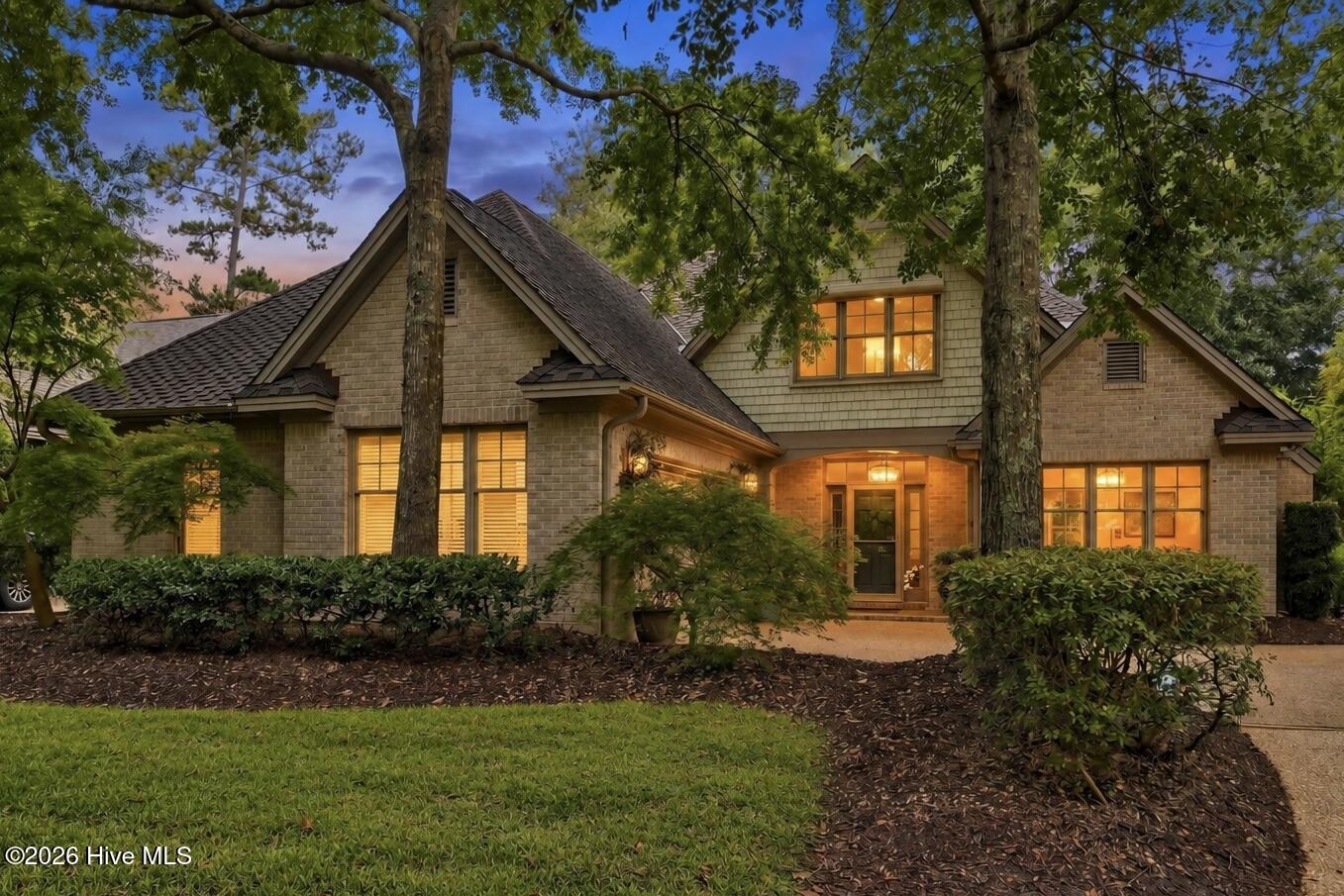 Two-story brick home with shingle gables and warmly lit windows, framed by mature trees at dusk.