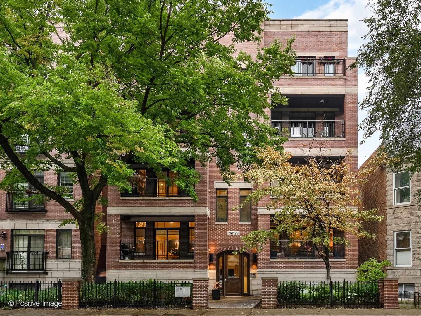 Brick apartment building with balconies, a gated entrance, and mature trees along the sidewalk.
