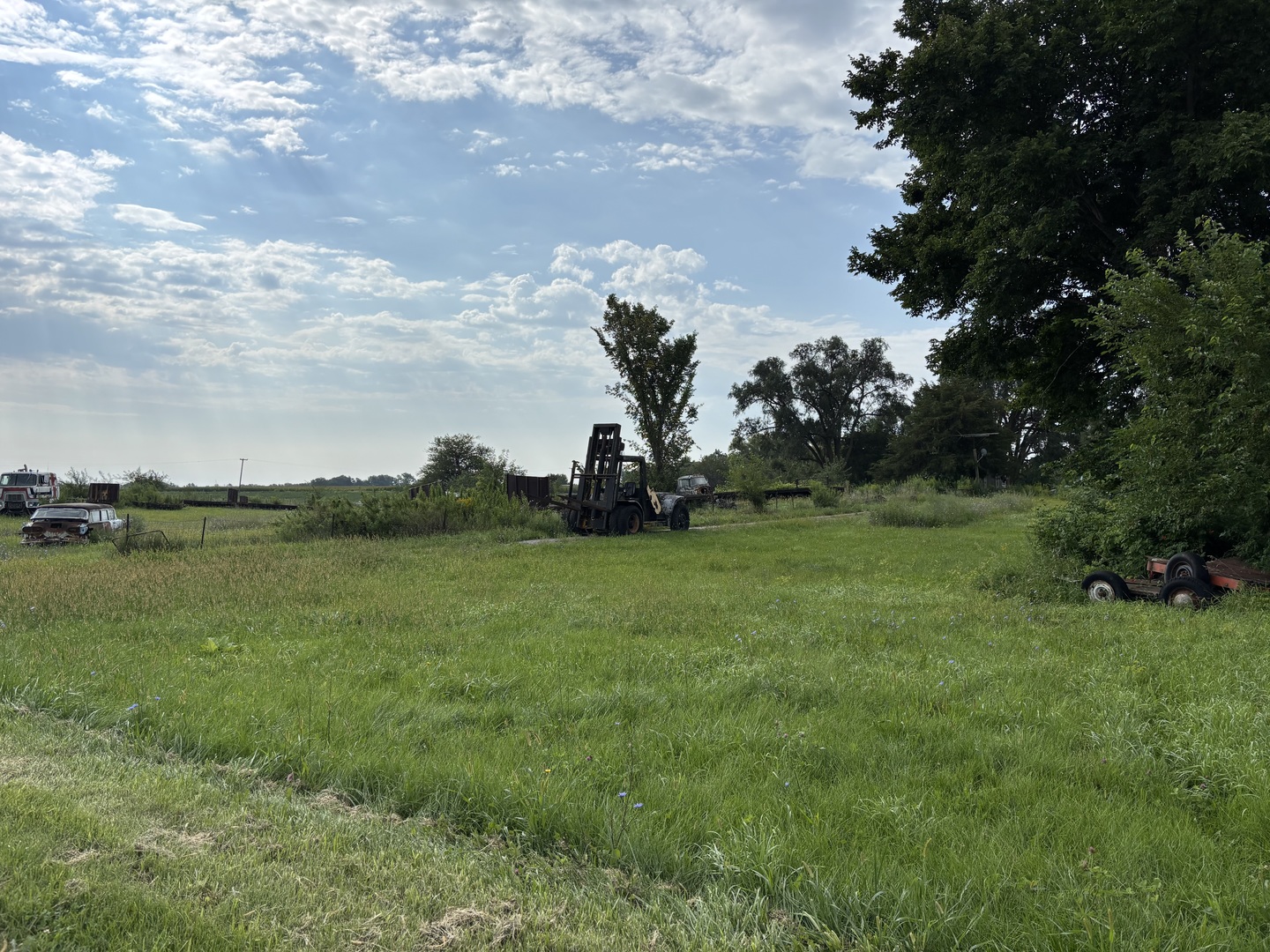 Grassy rural property with mature trees, a forklift, and old vehicles and trailers.