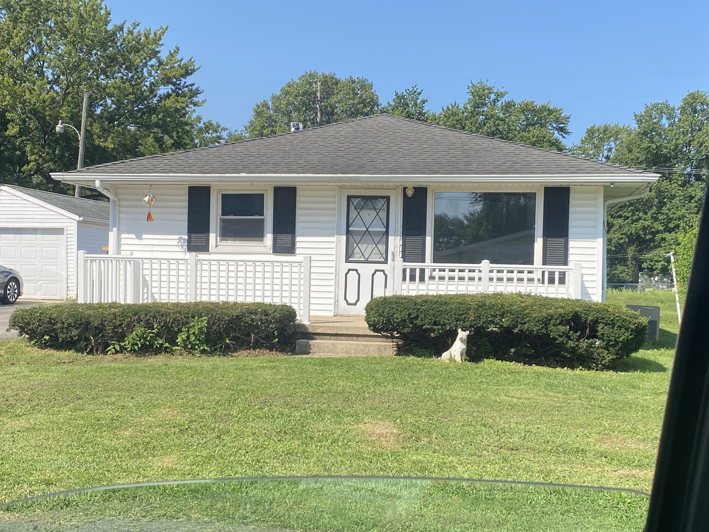 White single-story home with a covered porch, dark shutters, trimmed hedges, and a small dog.