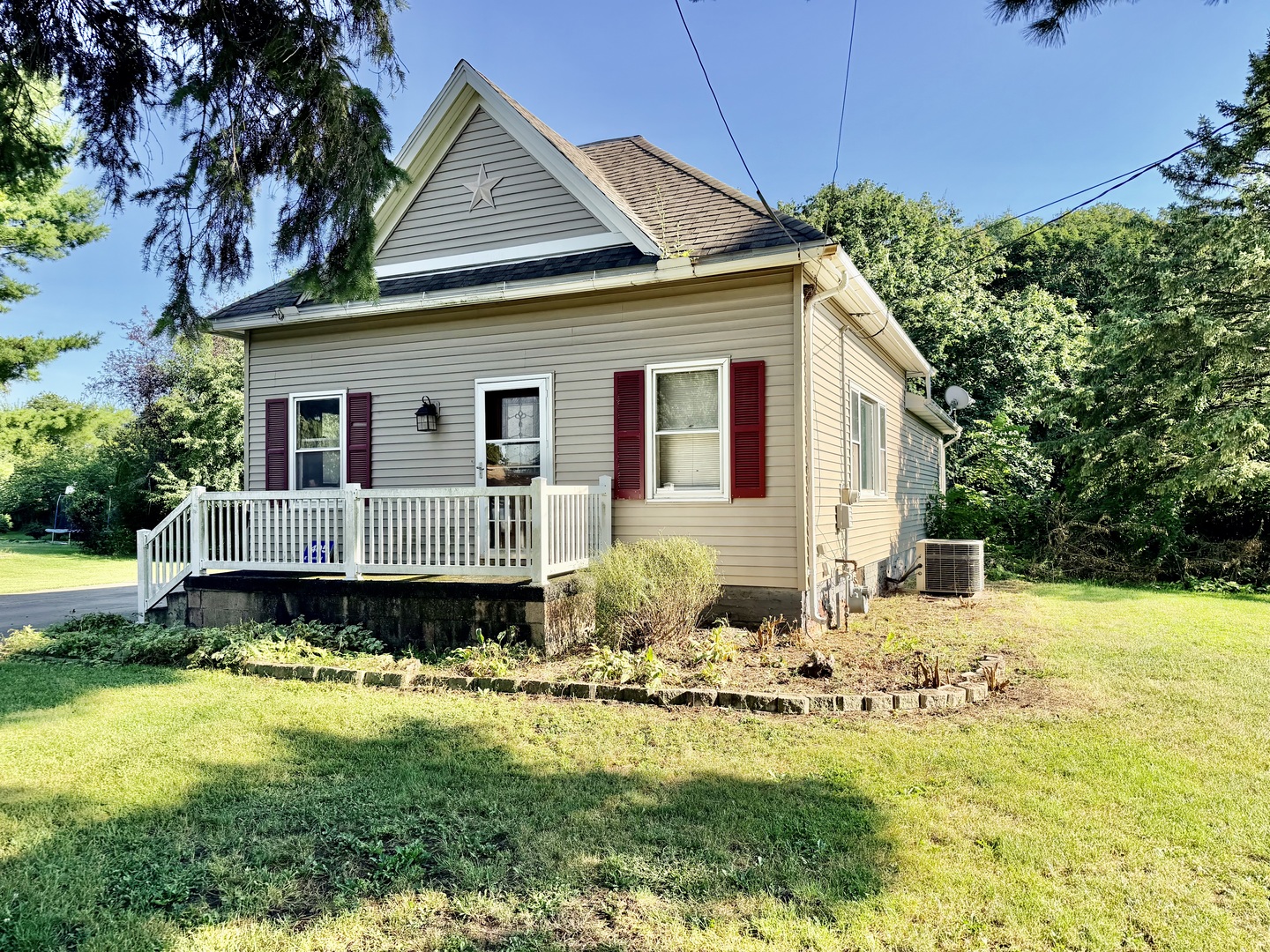 Beige house with burgundy shutters and a white-railed front porch beneath mature trees.