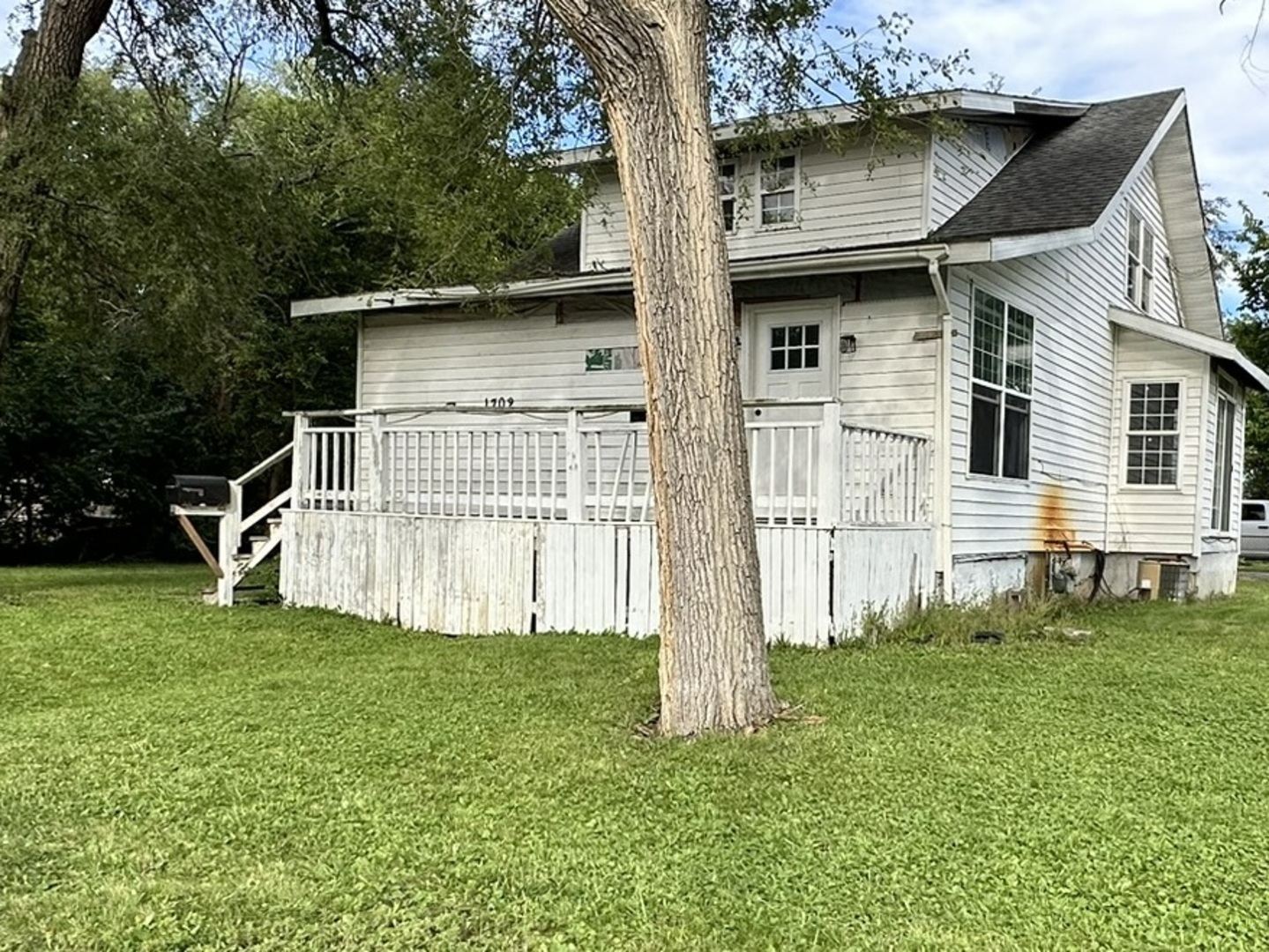 White-sided house with a raised porch, partly obscured by a large tree in the front lawn.