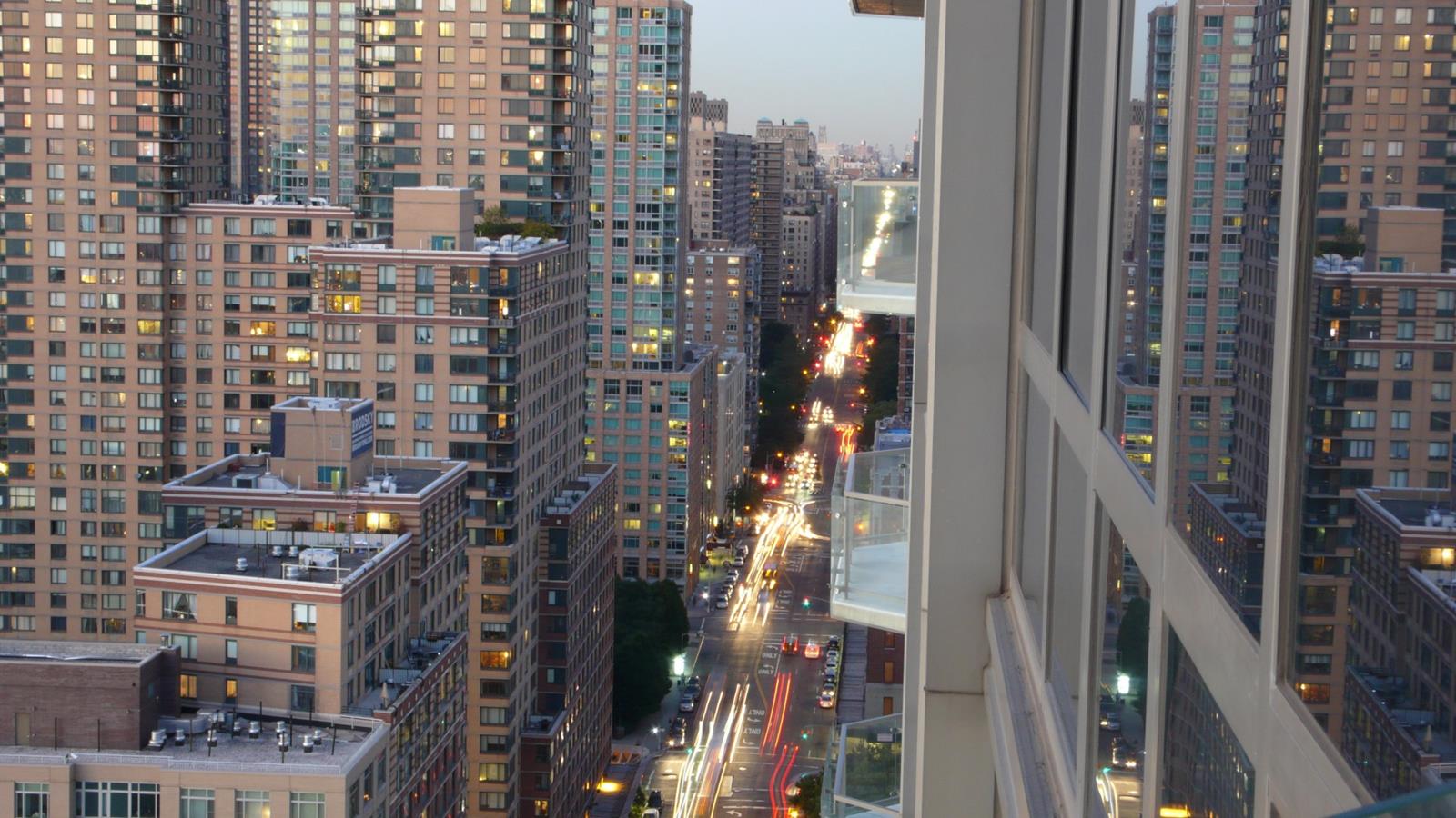Dusk city view from an apartment window, with high-rises and traffic light trails below.