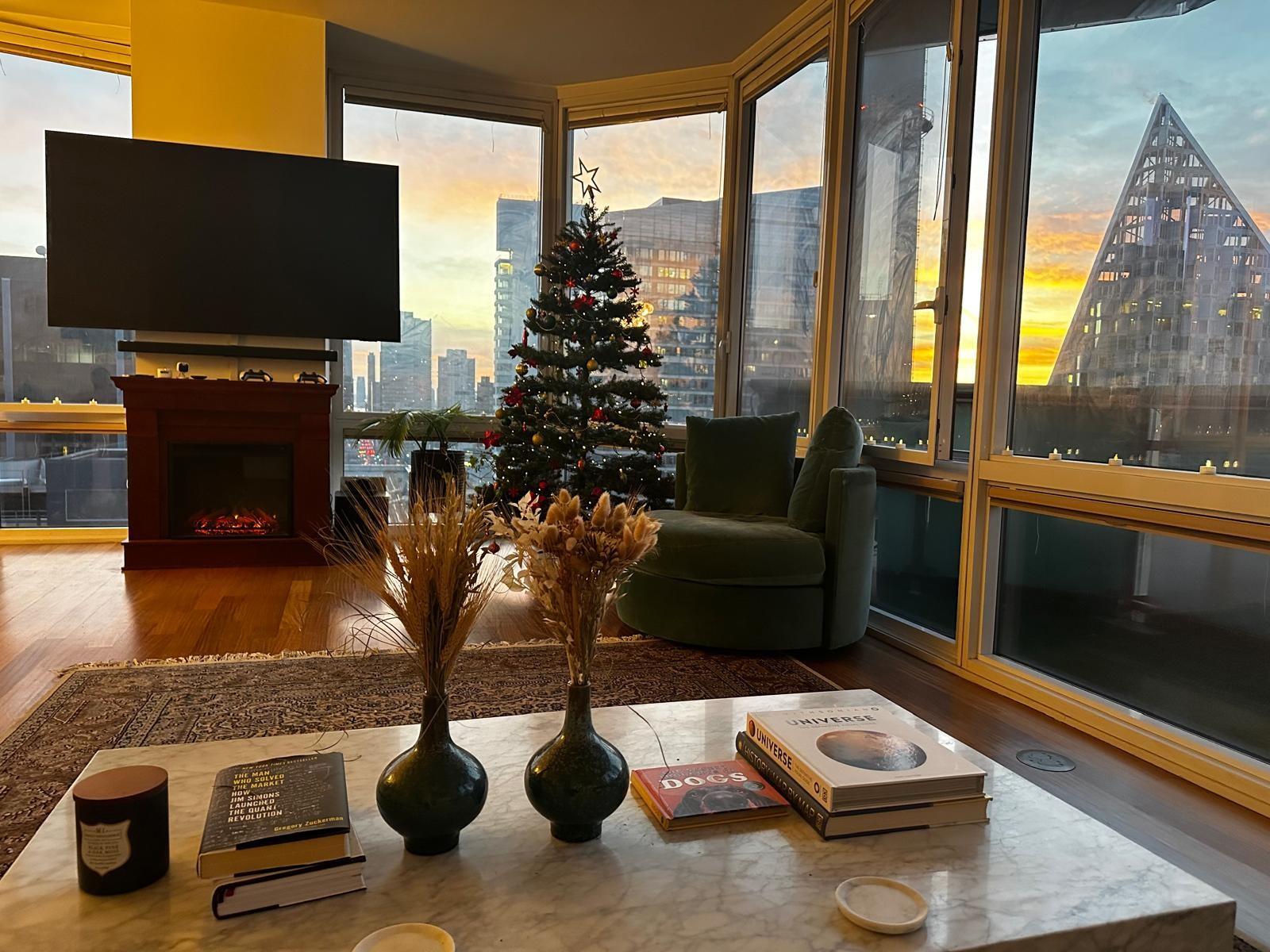 Living room with a Christmas tree, corner windows, and a city skyline at sunset.