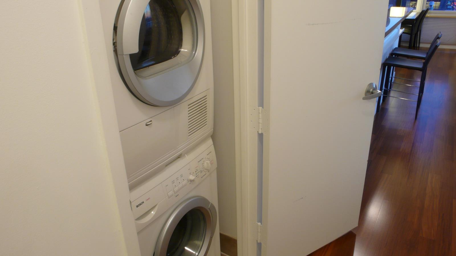 Stacked washer and dryer in a closet beside an open door to a hardwood-floored room.
