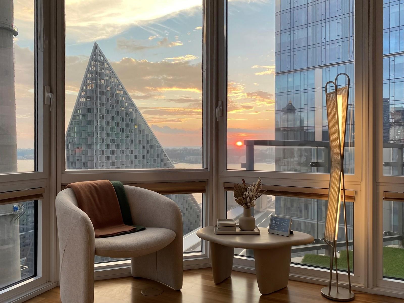 Furnished sitting area with floor-to-ceiling windows overlooking the skyline and river at sunset.