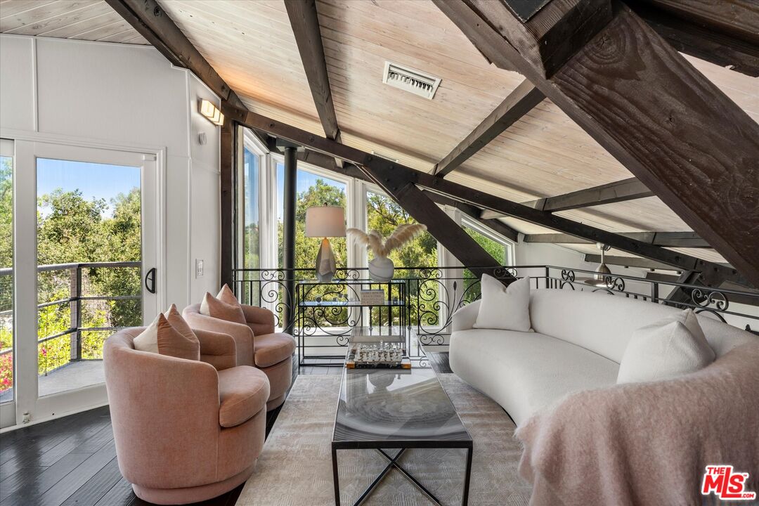 Loft sitting area with exposed timber beams, blush armchairs, and tall windows overlooking greenery.