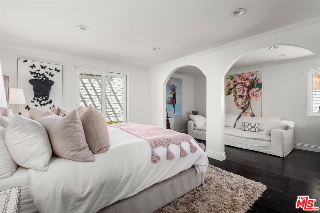 Furnished bedroom with a white bed, pink throw, and adjoining sitting area through arched openings.