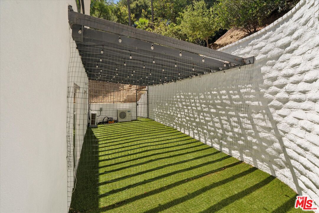 Enclosed artificial-turf side yard beneath a pergola with string lights and white masonry walls.