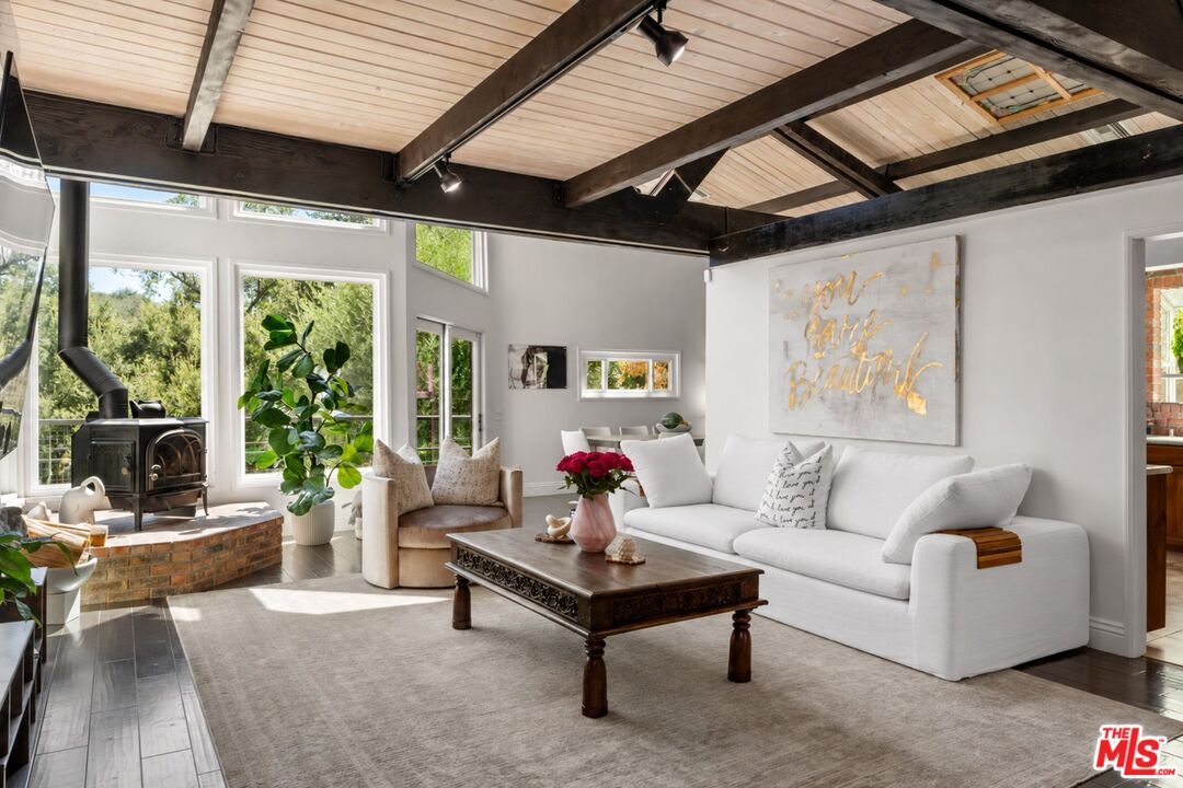 Living room with exposed beams, large windows, a white sofa, and a freestanding wood stove.