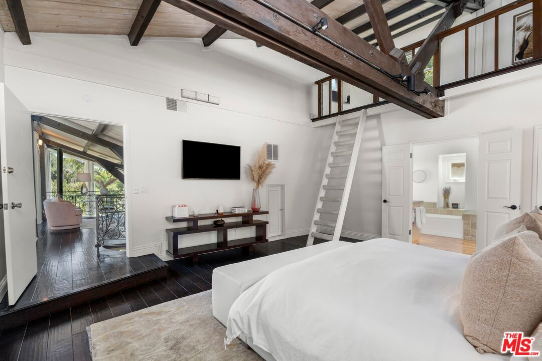 Bedroom with exposed timber beams, a loft ladder, and doors to a bathroom and covered balcony.