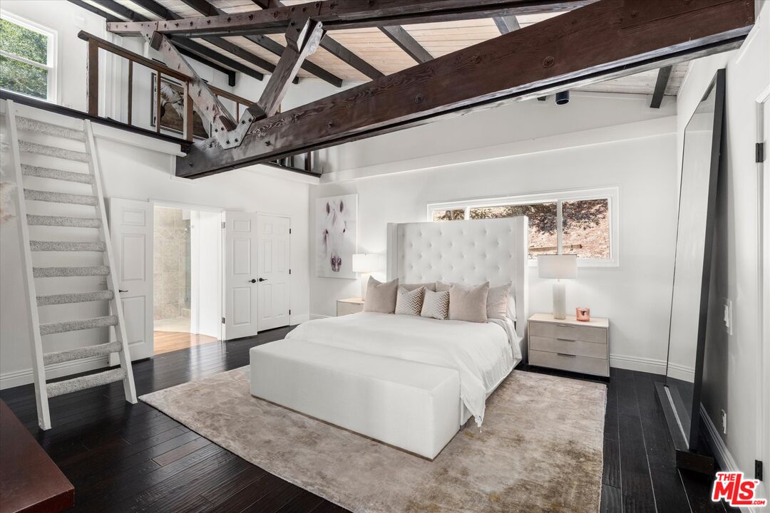 Staged bedroom with a white upholstered bed, exposed timber beams, and a staircase to the loft.