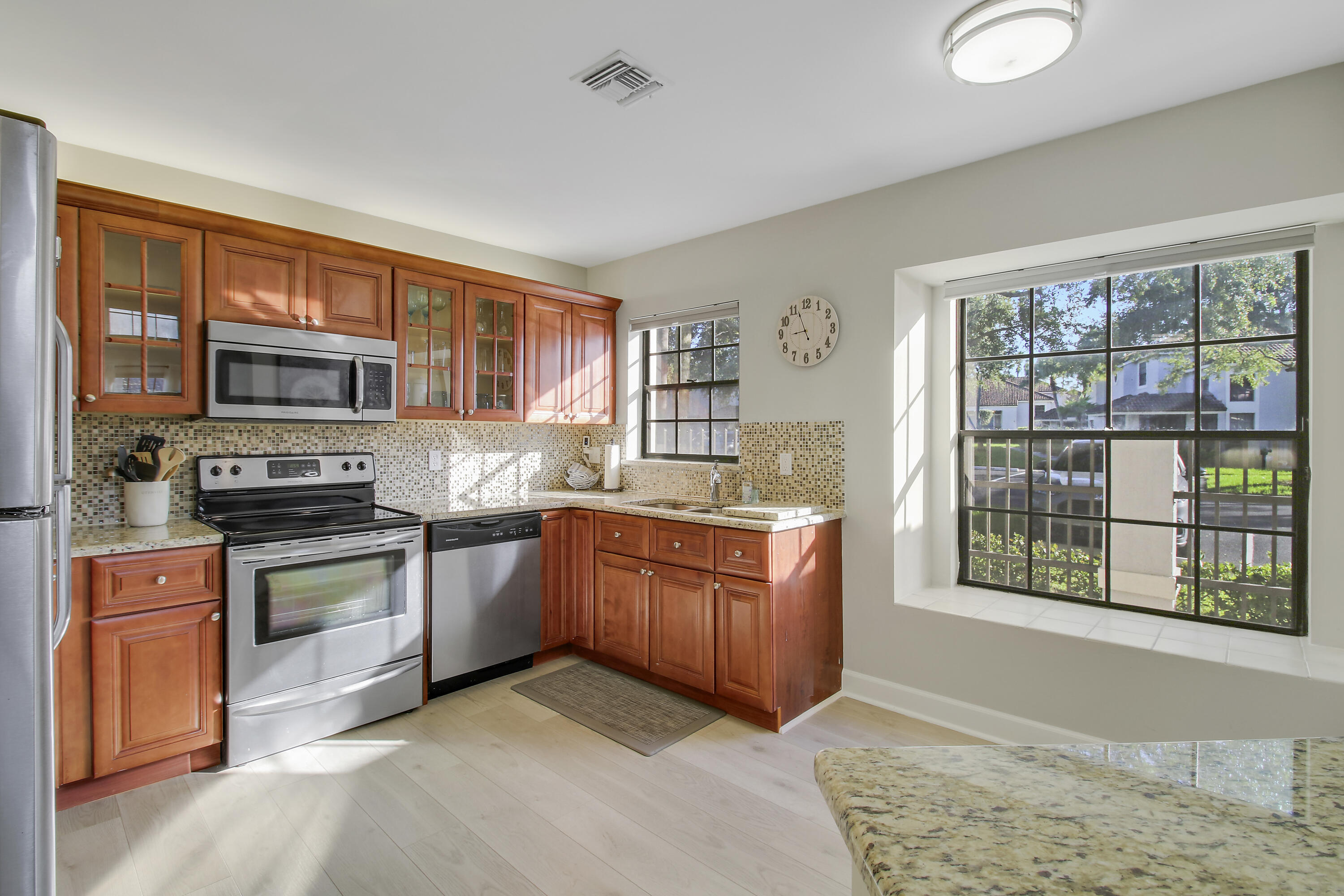 Kitchen with reddish-brown cabinets, stainless steel appliances, mosaic backsplash, and large sunlit windows.