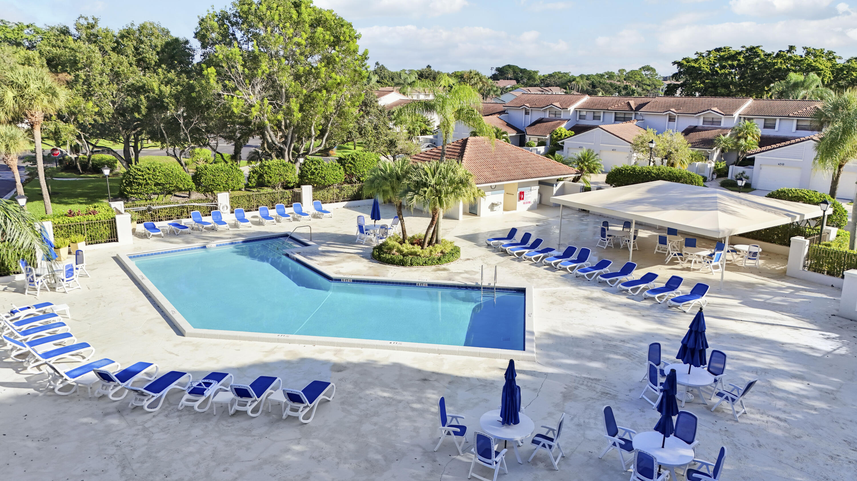 Community pool with blue lounge chairs, shaded tables, and palm trees beside nearby townhomes.