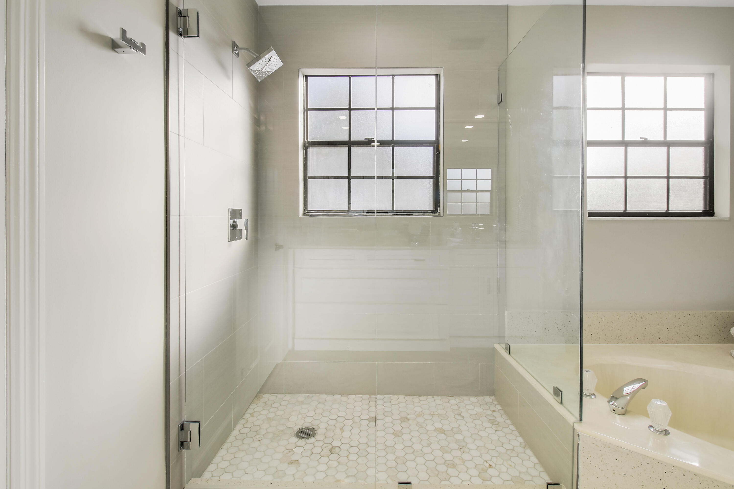 Modern bathroom with a glass walk-in shower, hexagonal tile floor, and built-in bathtub beside windows.