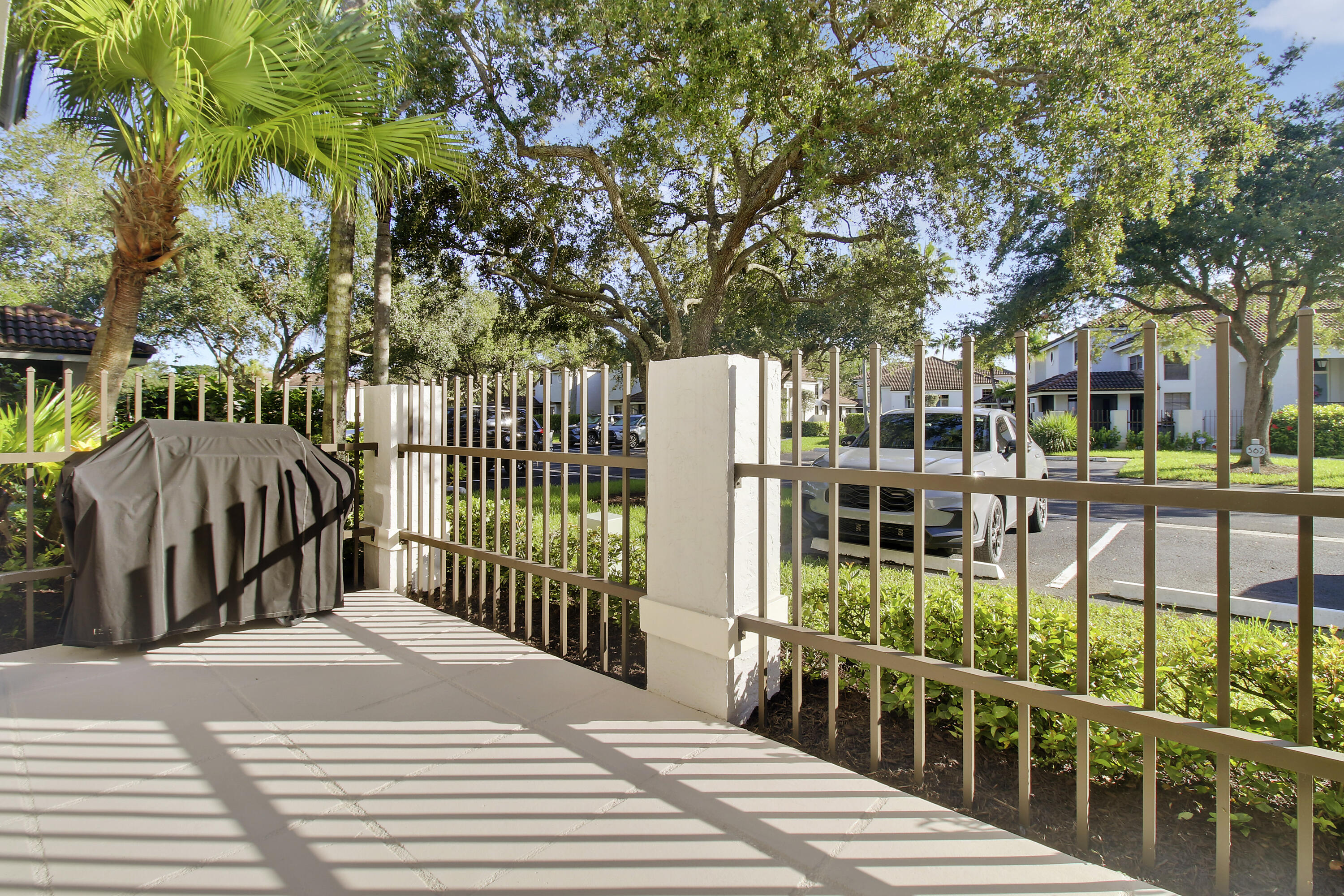 Paved patio with a covered grill, metal railing, and leafy trees overlooking a parking area.