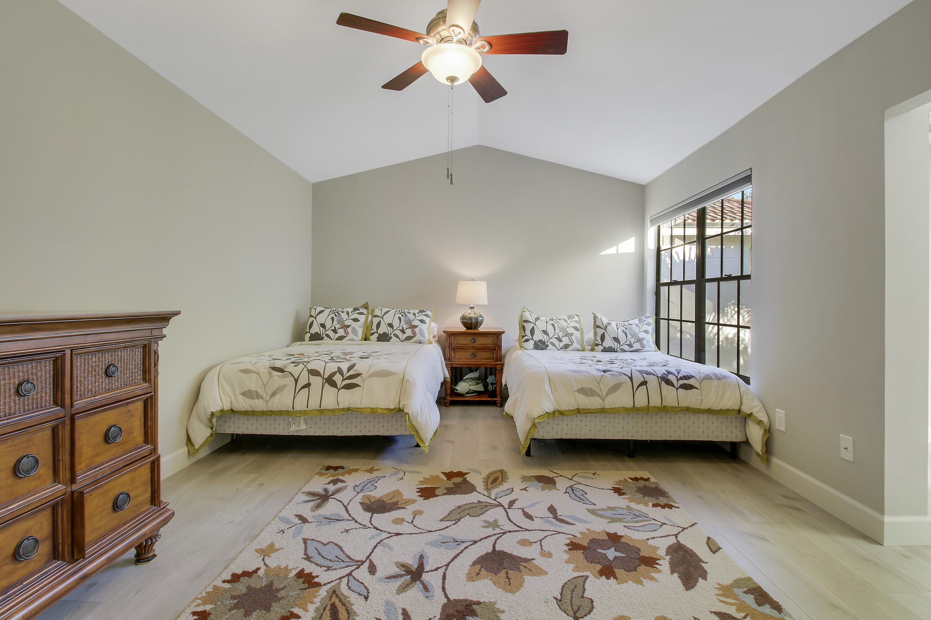 Furnished bedroom with two matching beds, a vaulted ceiling, and a large black-framed window.