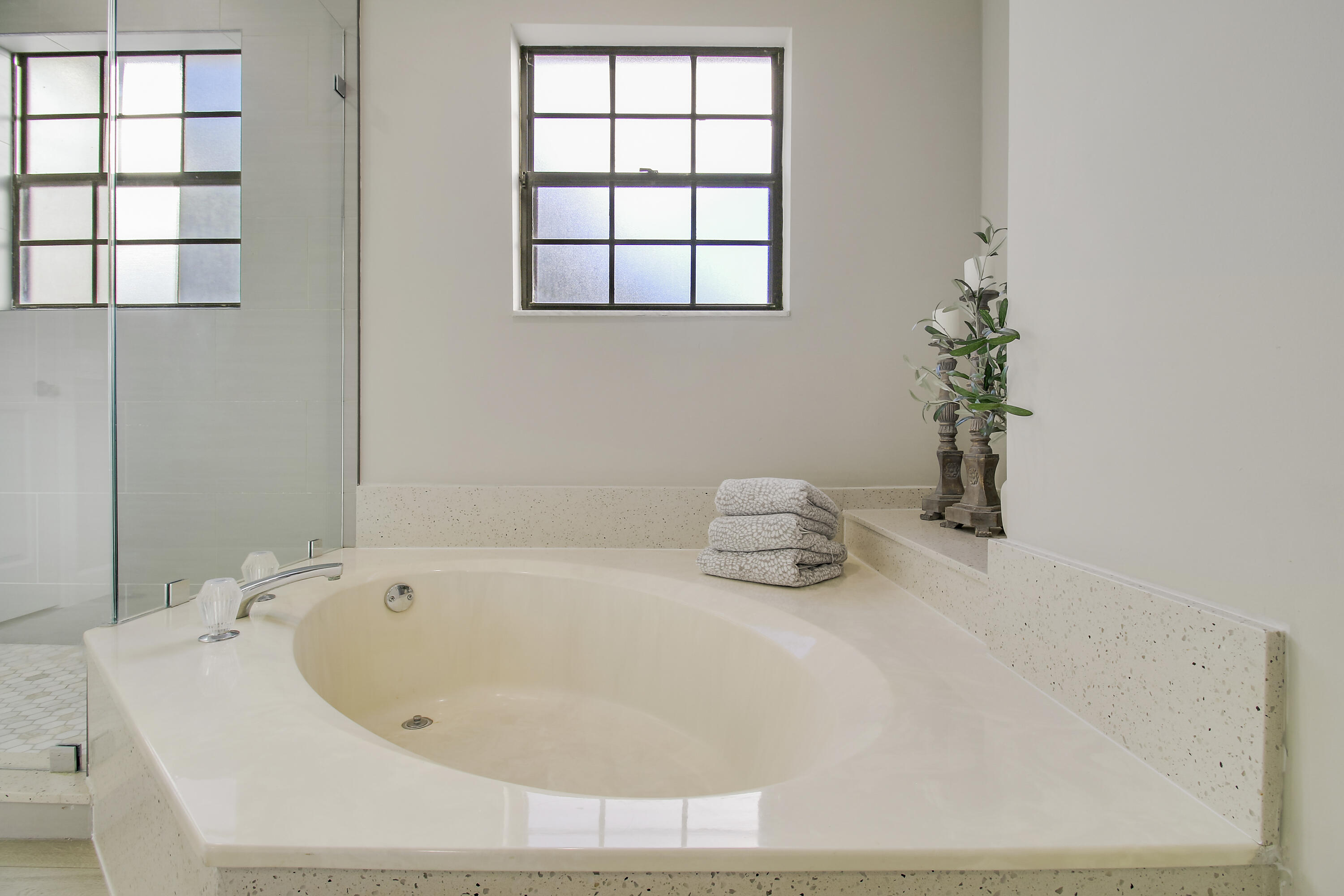 Built-in soaking tub with a speckled stone surround beside a glass shower and black-grid window.